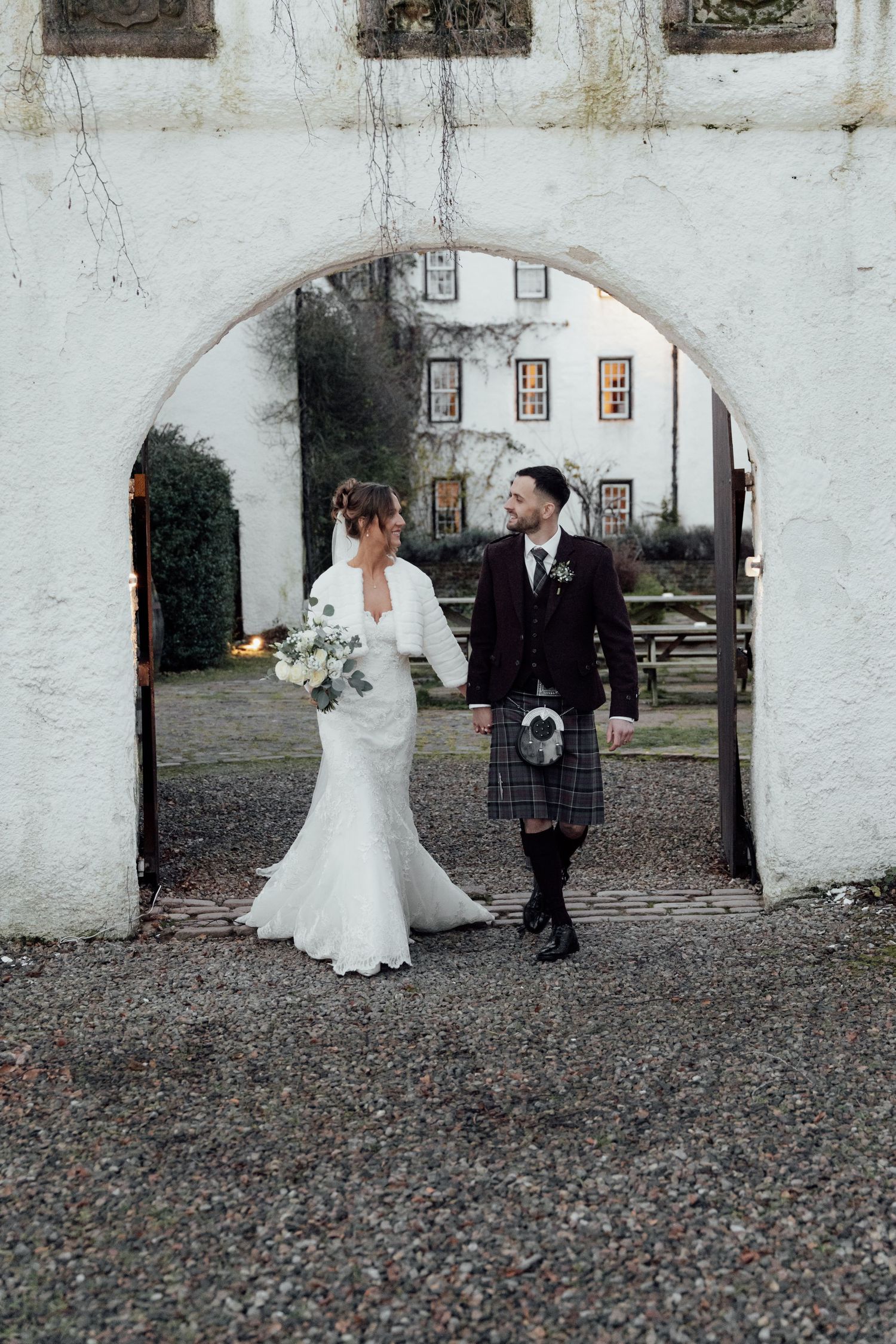 Wedding couple standing under white stone archway, bride in lace gown and groom in traditional Highland dress.