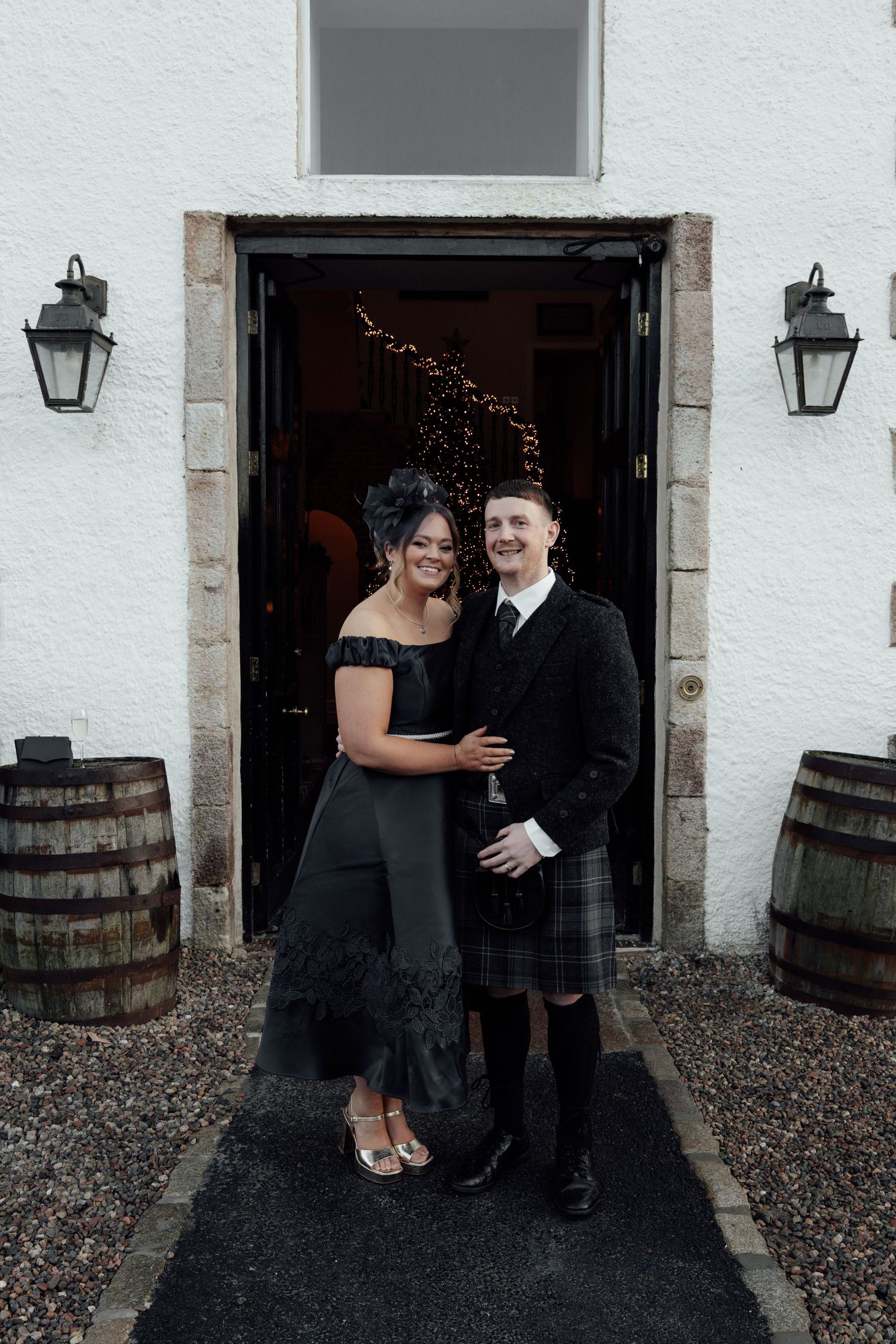 Joyful wedding party members pose casually against white stone wall with rustic barrels.