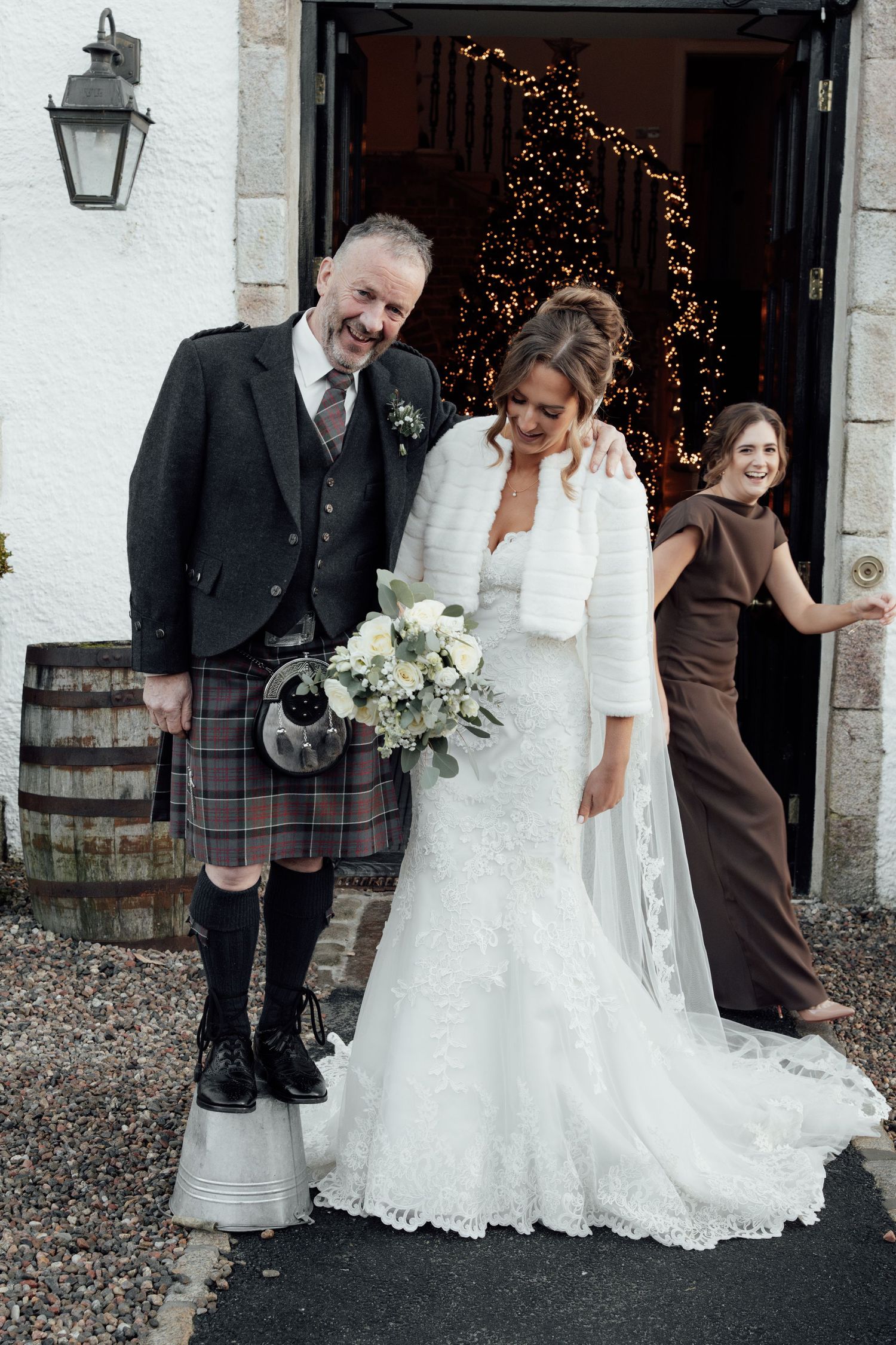 A wedding party poses in front of a white building with fairy lights and wooden barrels.