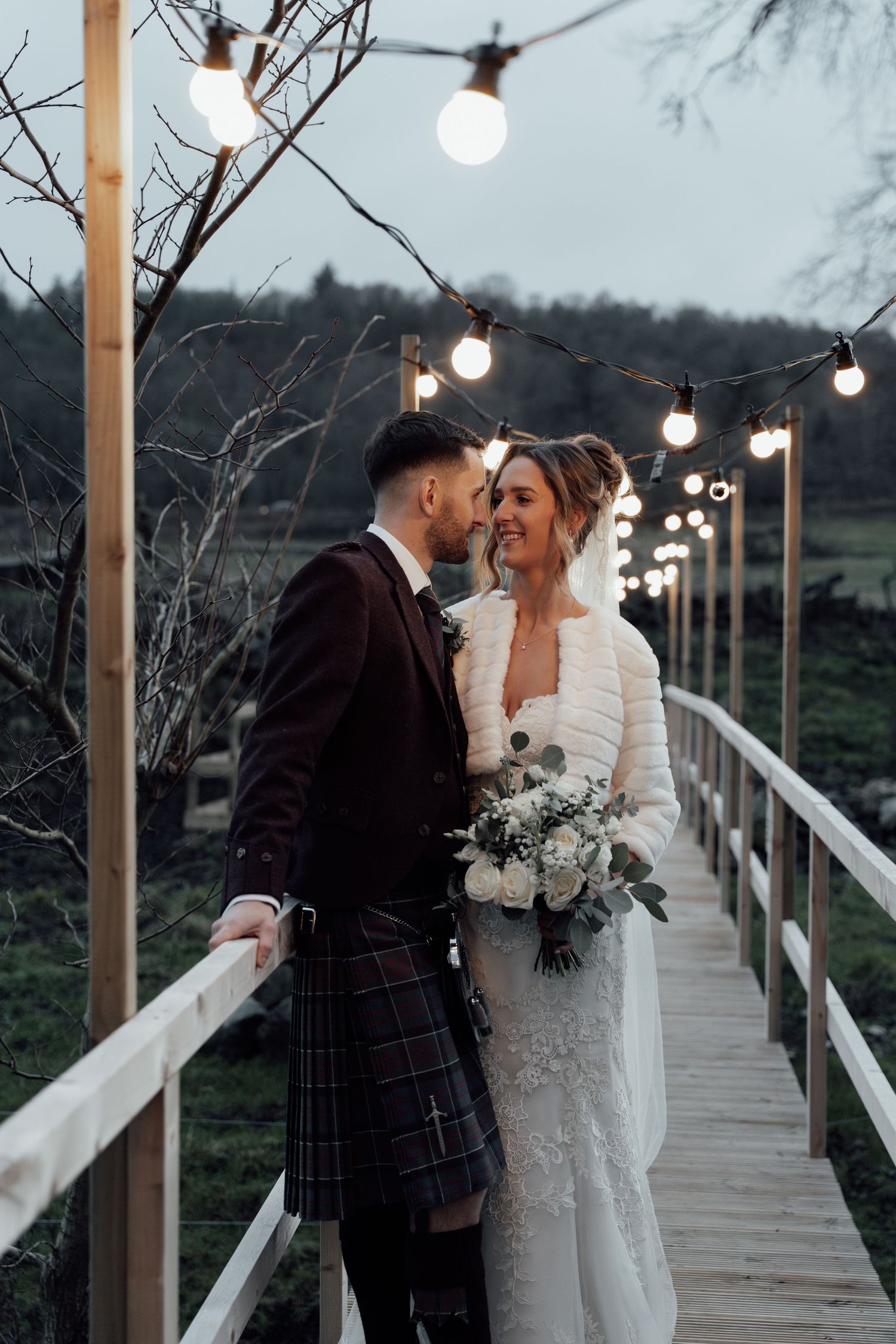 Wedding couple shares tender moment on wooden bridge with string lights and winter scenery.
