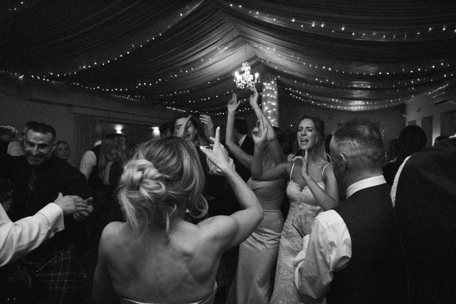 Wedding reception dance floor filled with guests under draped ceiling and string lights.