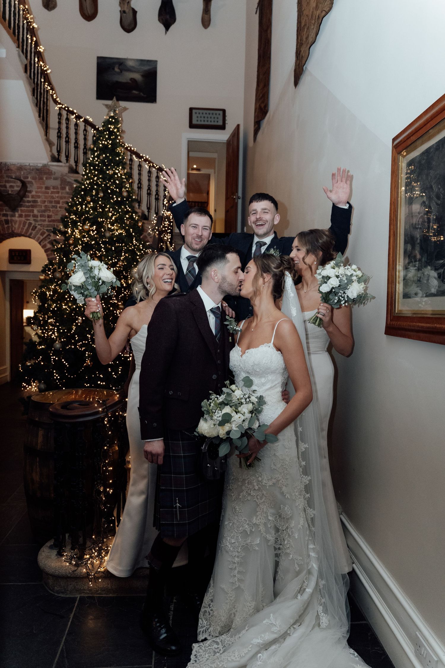 Wedding celebration with festive Christmas tree and joyful group waving in indoor hallway.