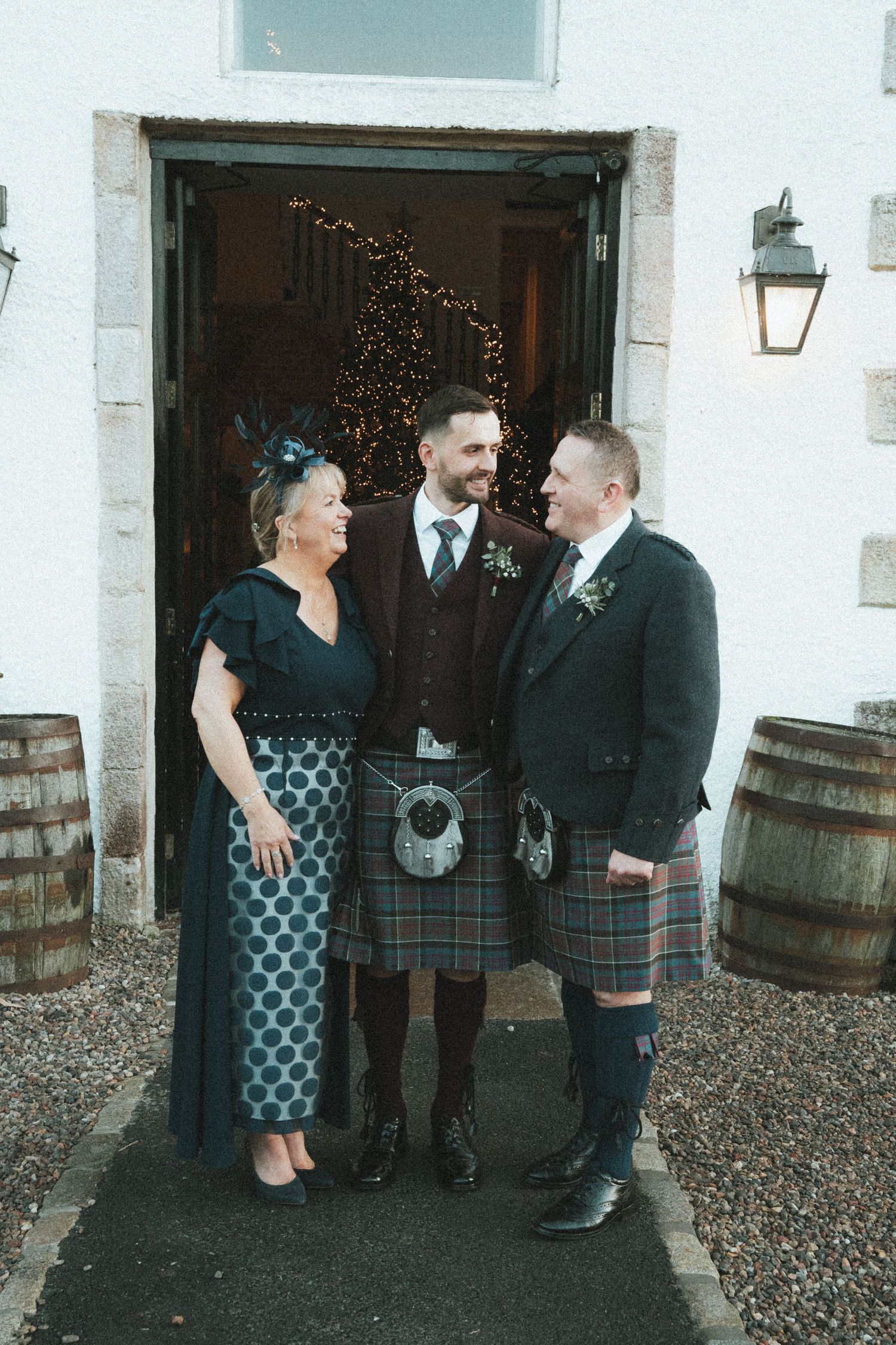 Wedding party members in kilts and formal wear gather for photos outside a venue.