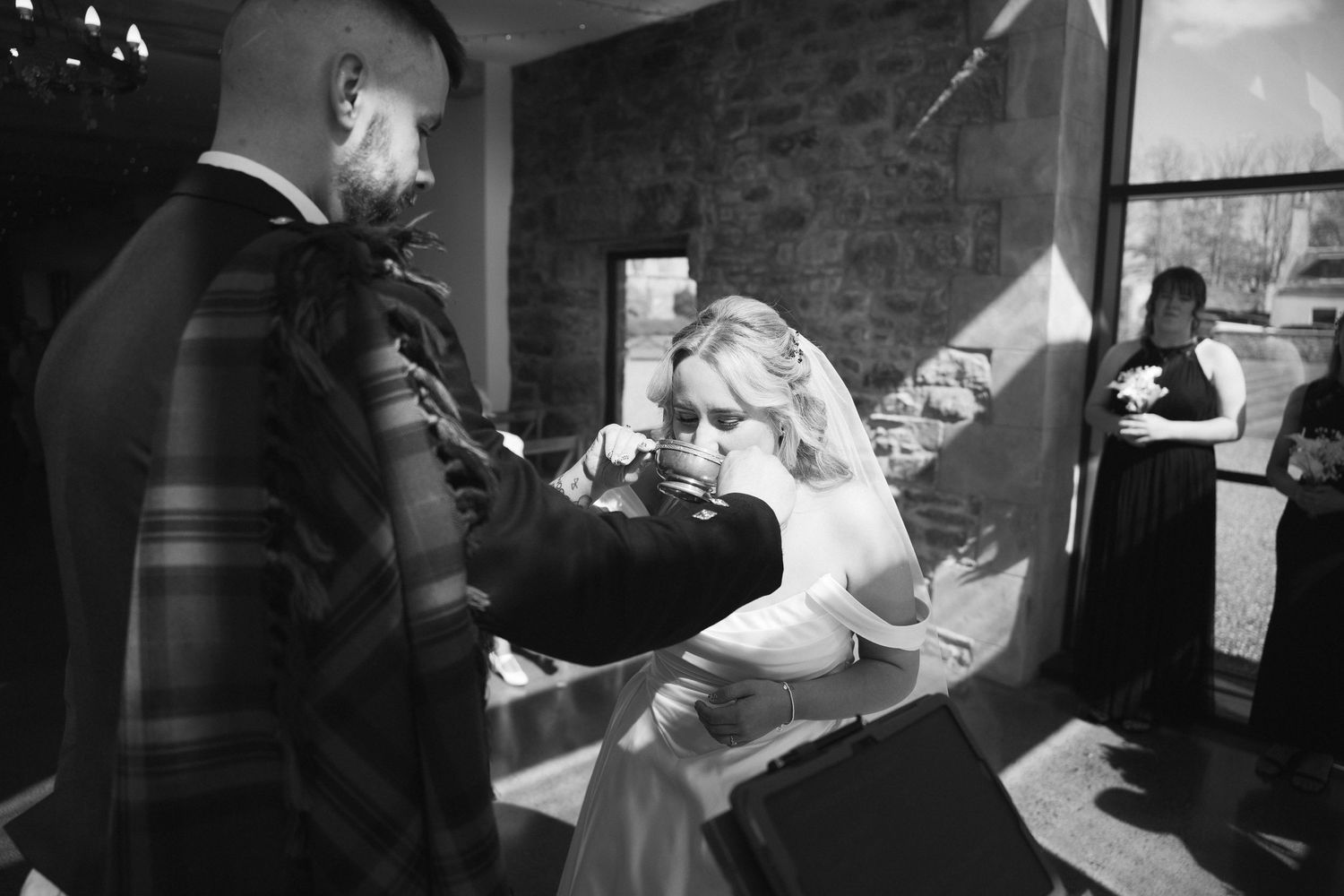 Black and white photo of groom in tartan kilt helping emotional bride during intimate wedding ceremony moment.