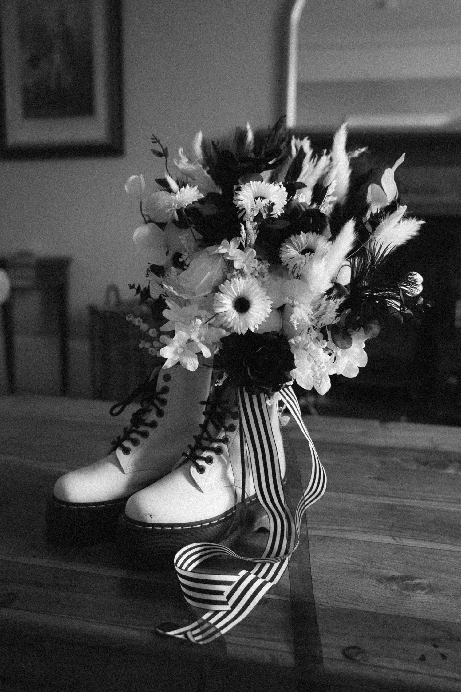 Black and white photo of bridal bouquet resting against Dr. Martens boots with striped ribbon on wooden table.