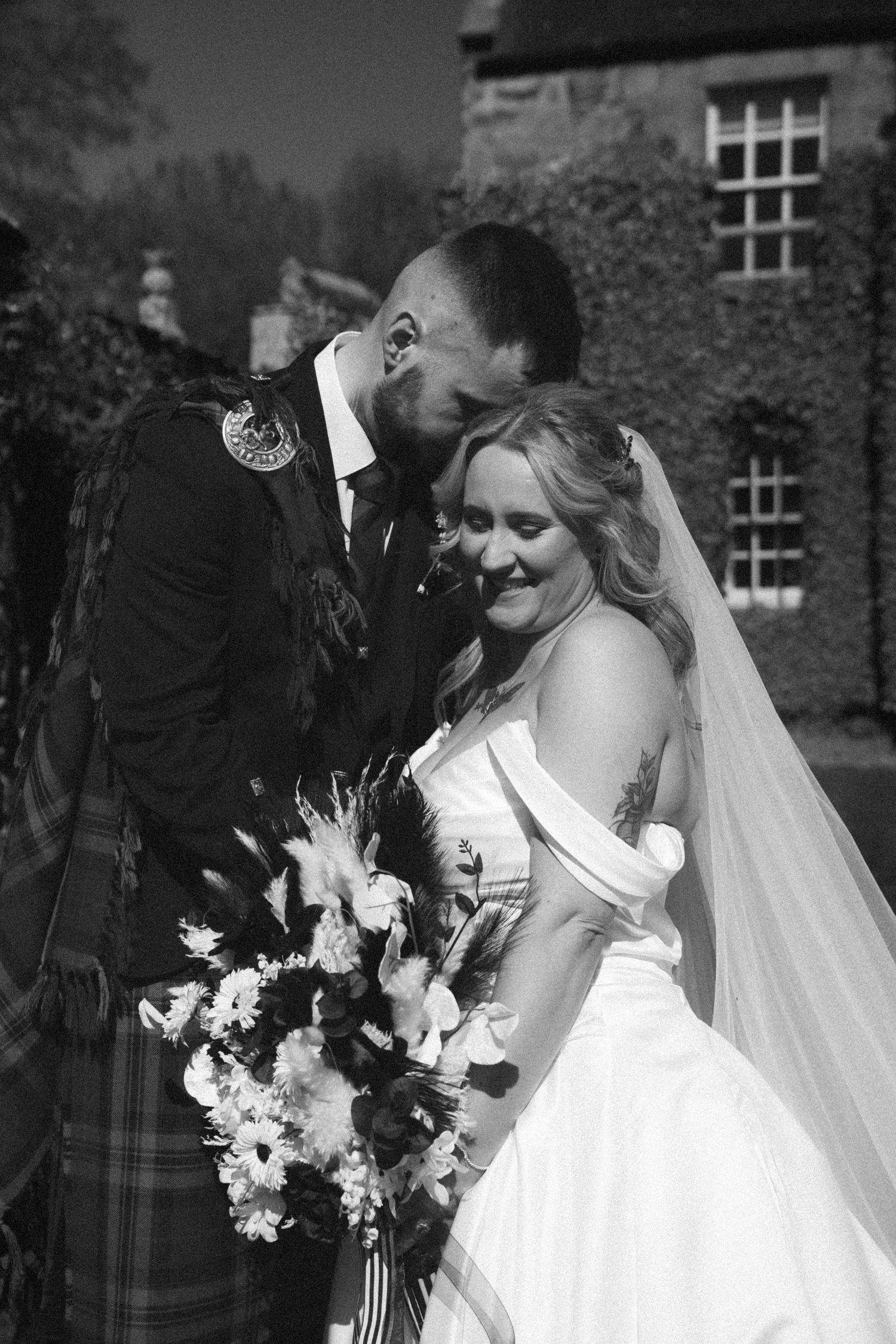 Black and white portrait of groom in tartan kilt embracing bride holding large floral bouquet outside stone manor.