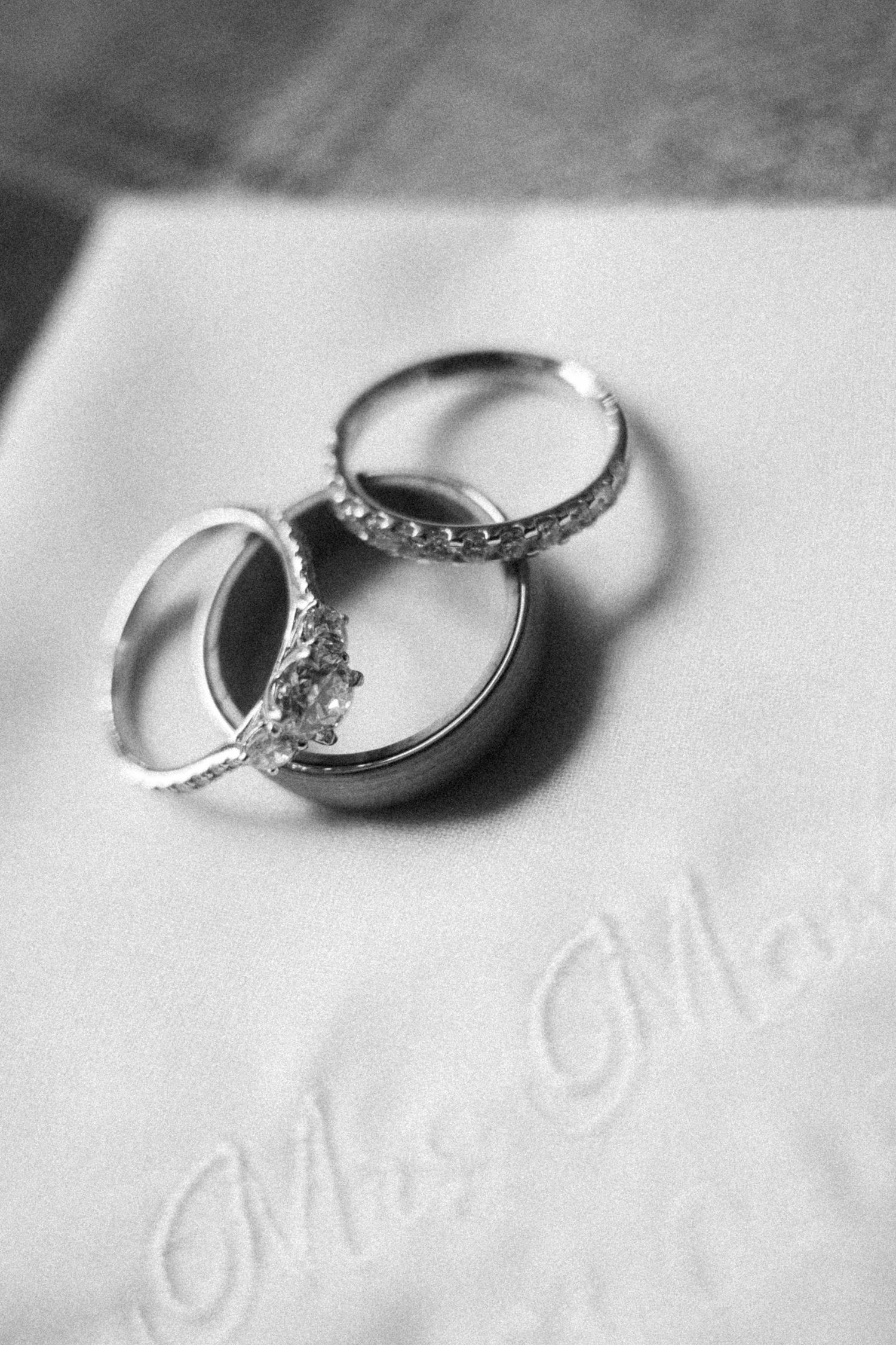 Black and white photo of wedding rings and engagement ring resting on embroidered Mrs. handkerchief.