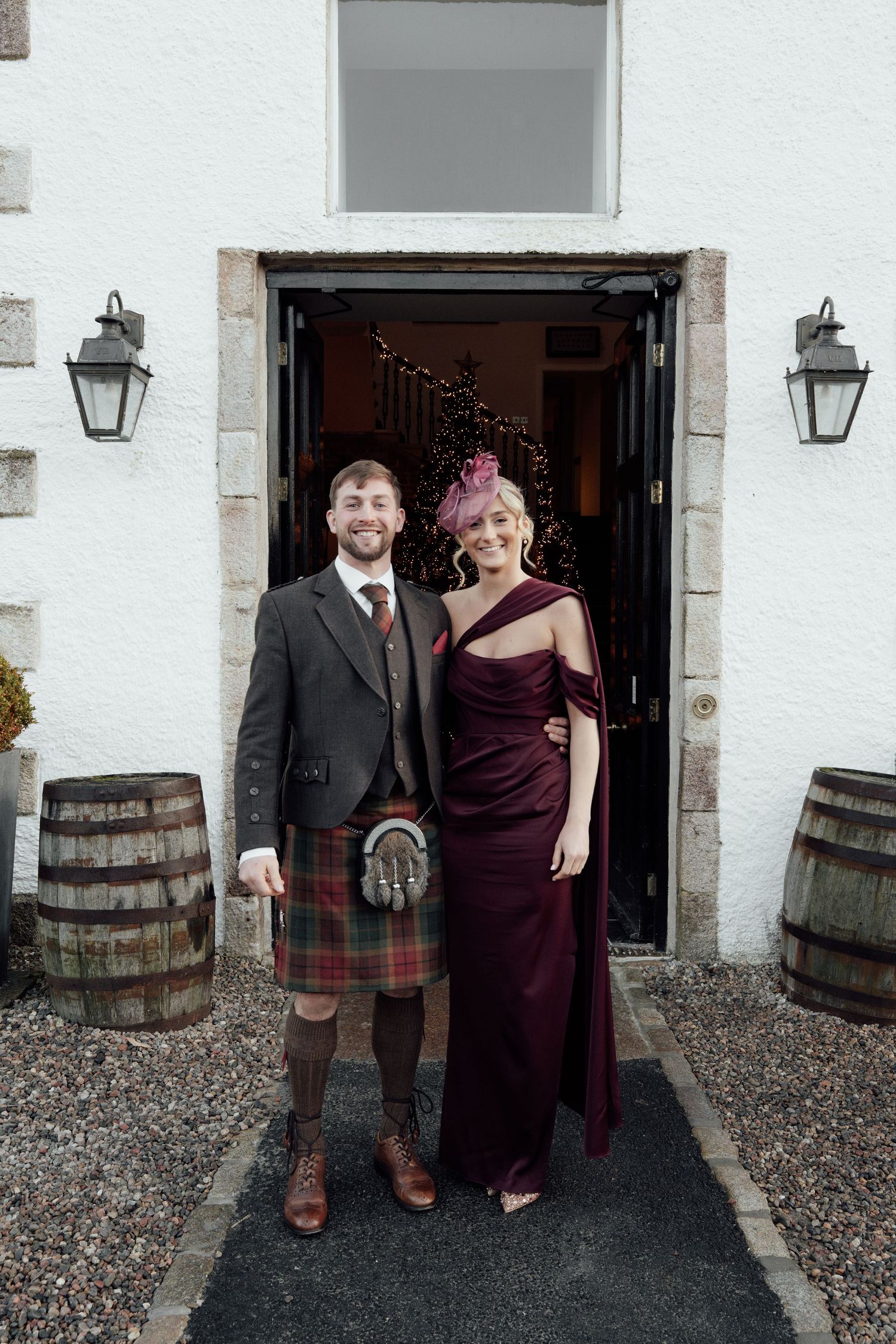 Wedding party members in burgundy dress and traditional Scottish kilt pose by wooden barrels.