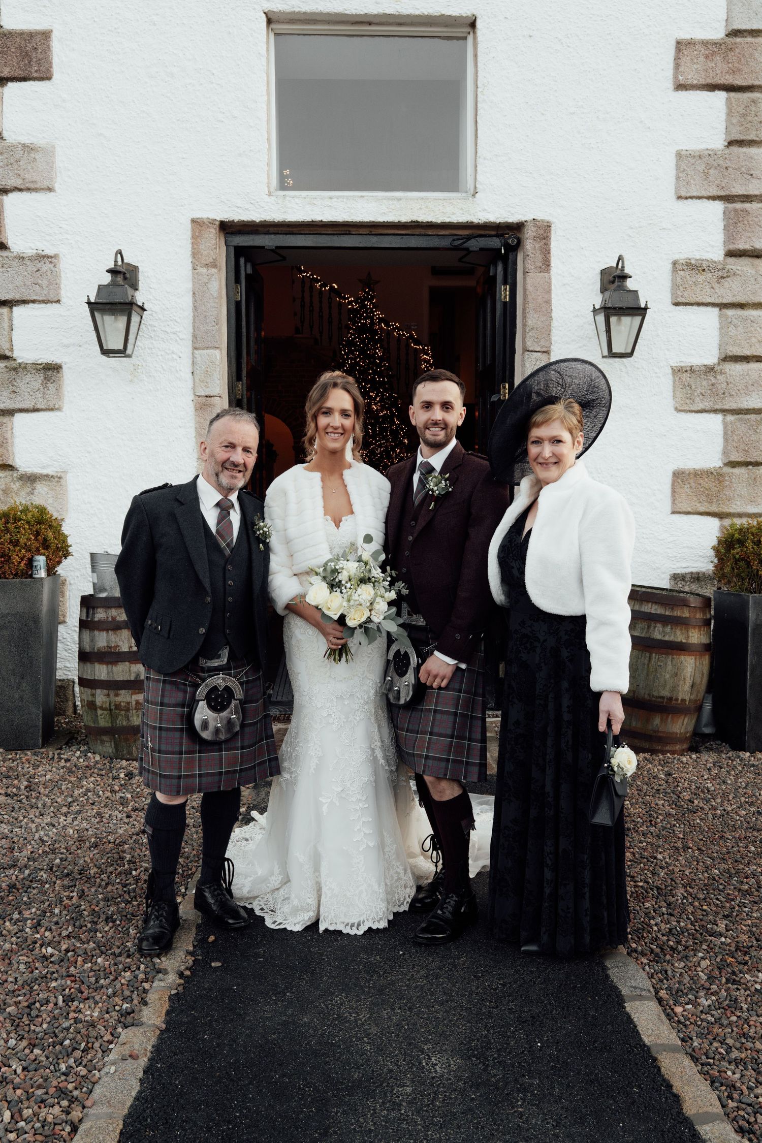 A wedding party of four poses formally outside a white building with traditional Scottish elements.