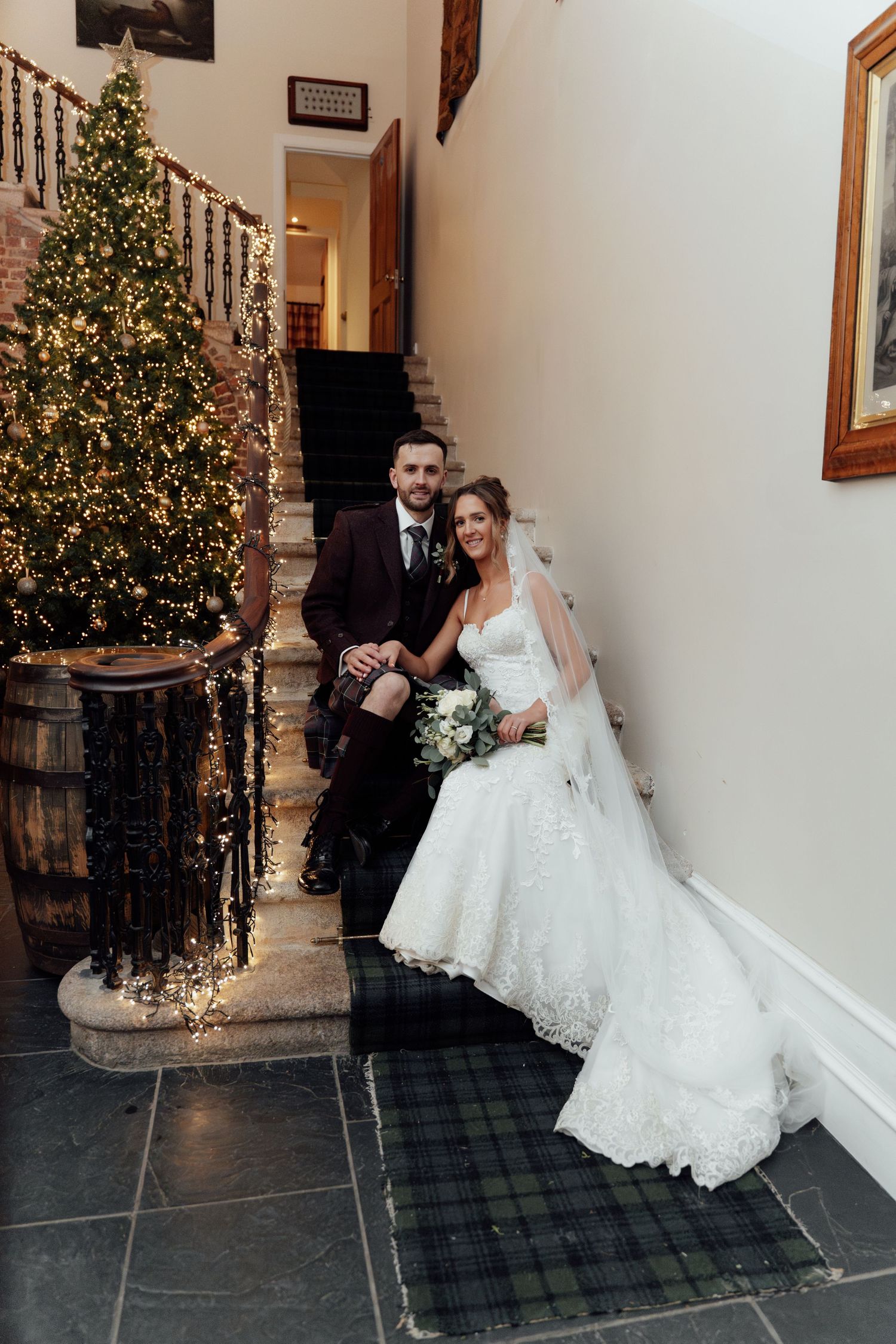 Wedding couple poses playfully by decorated Christmas tree in elegant indoor venue.