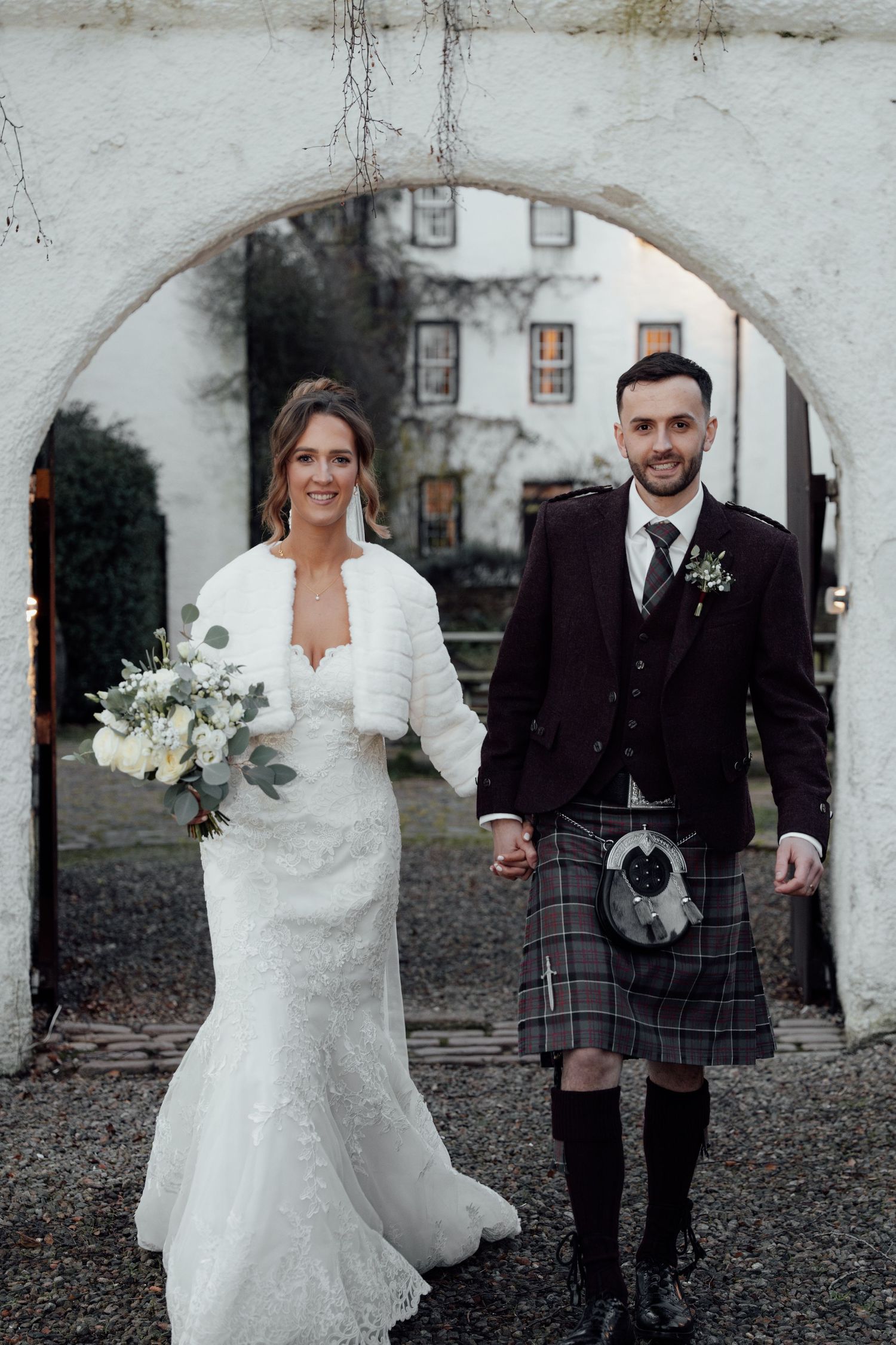 Elegant wedding portrait under archway with bride in lace gown and groom in traditional Scottish formal wear.