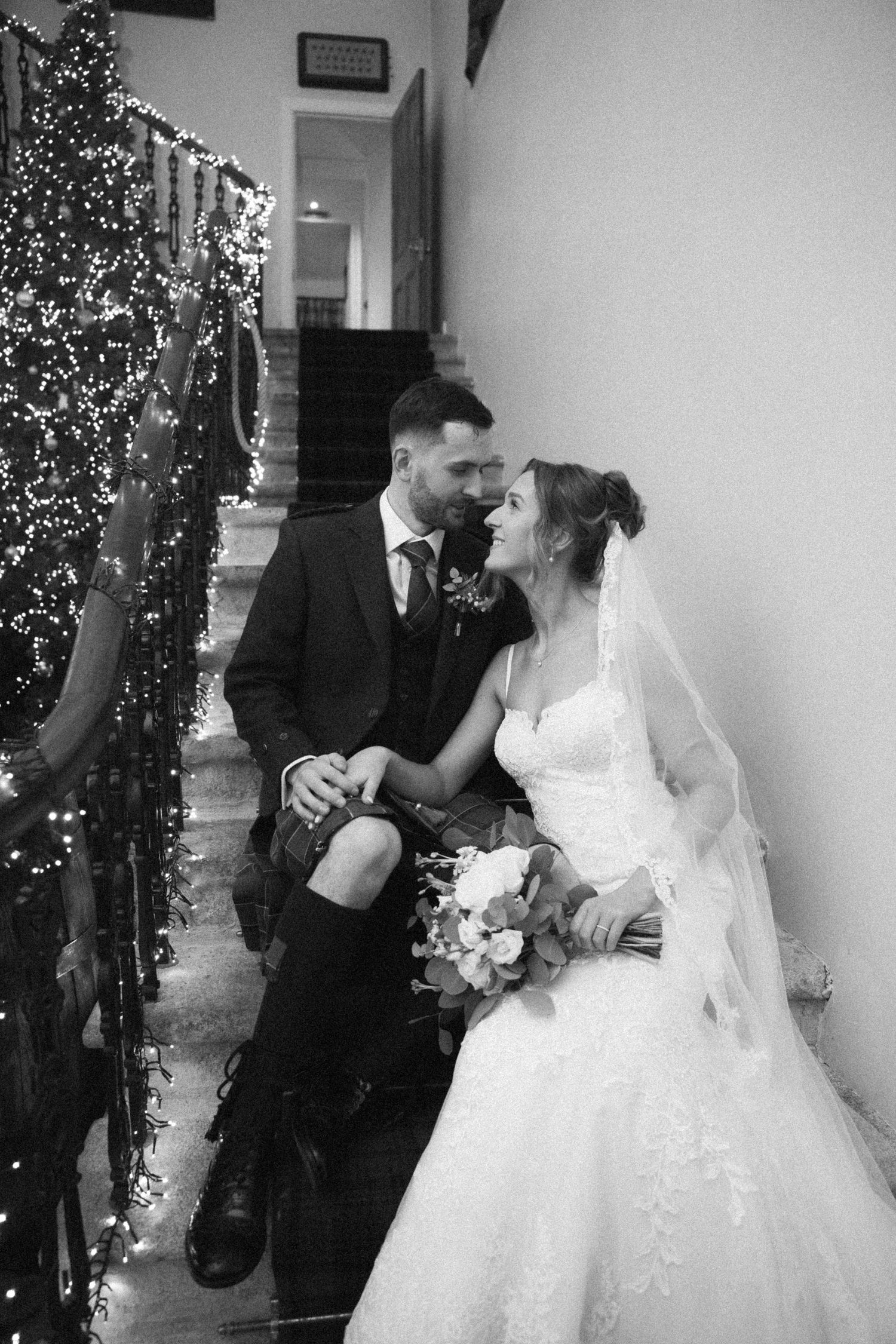 Black and white wedding photo of couple seated on stairs with Christmas lights and decorations.