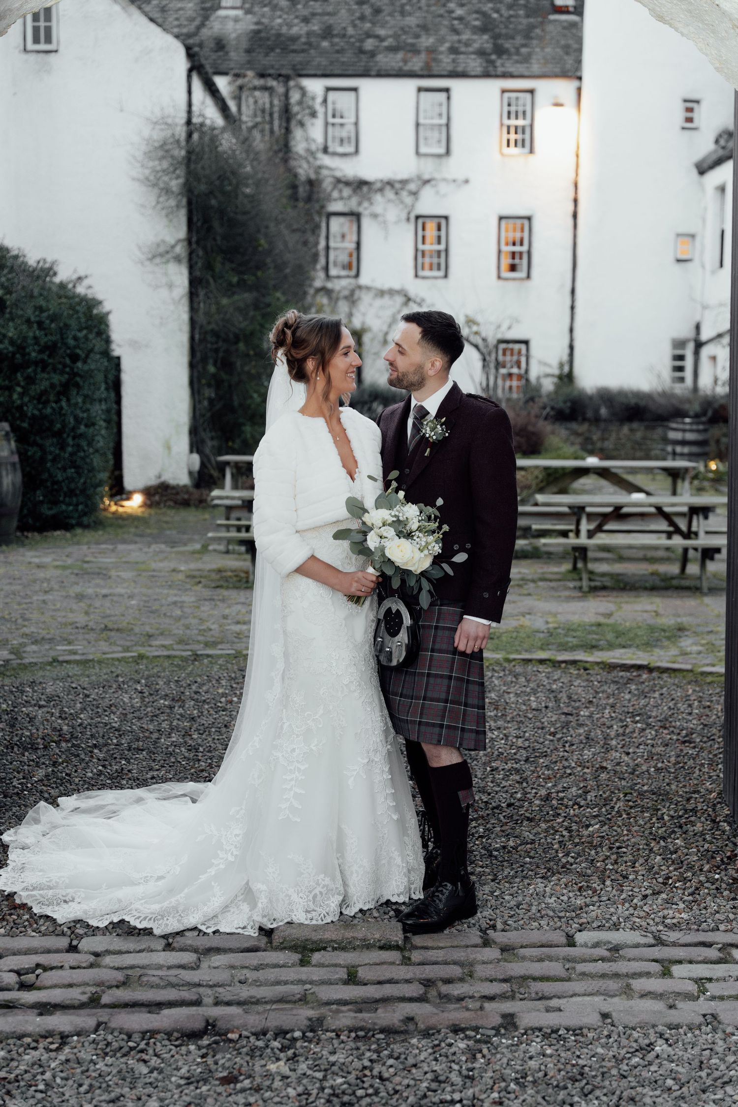 Wedding portrait of couple in Scottish attire against historic building backdrop.
