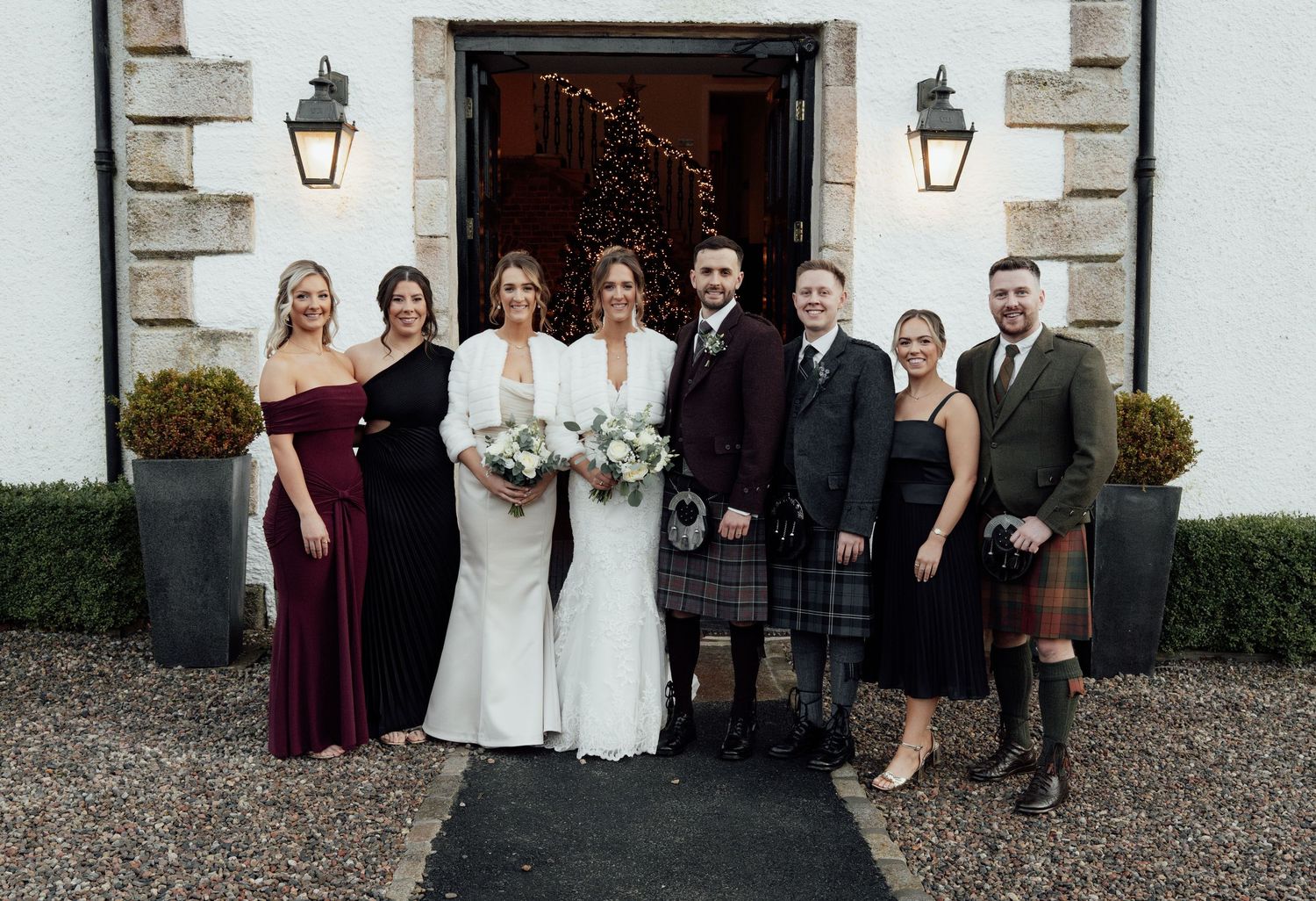 Wedding party poses for formal photos outside a white stone venue with decorative lanterns.