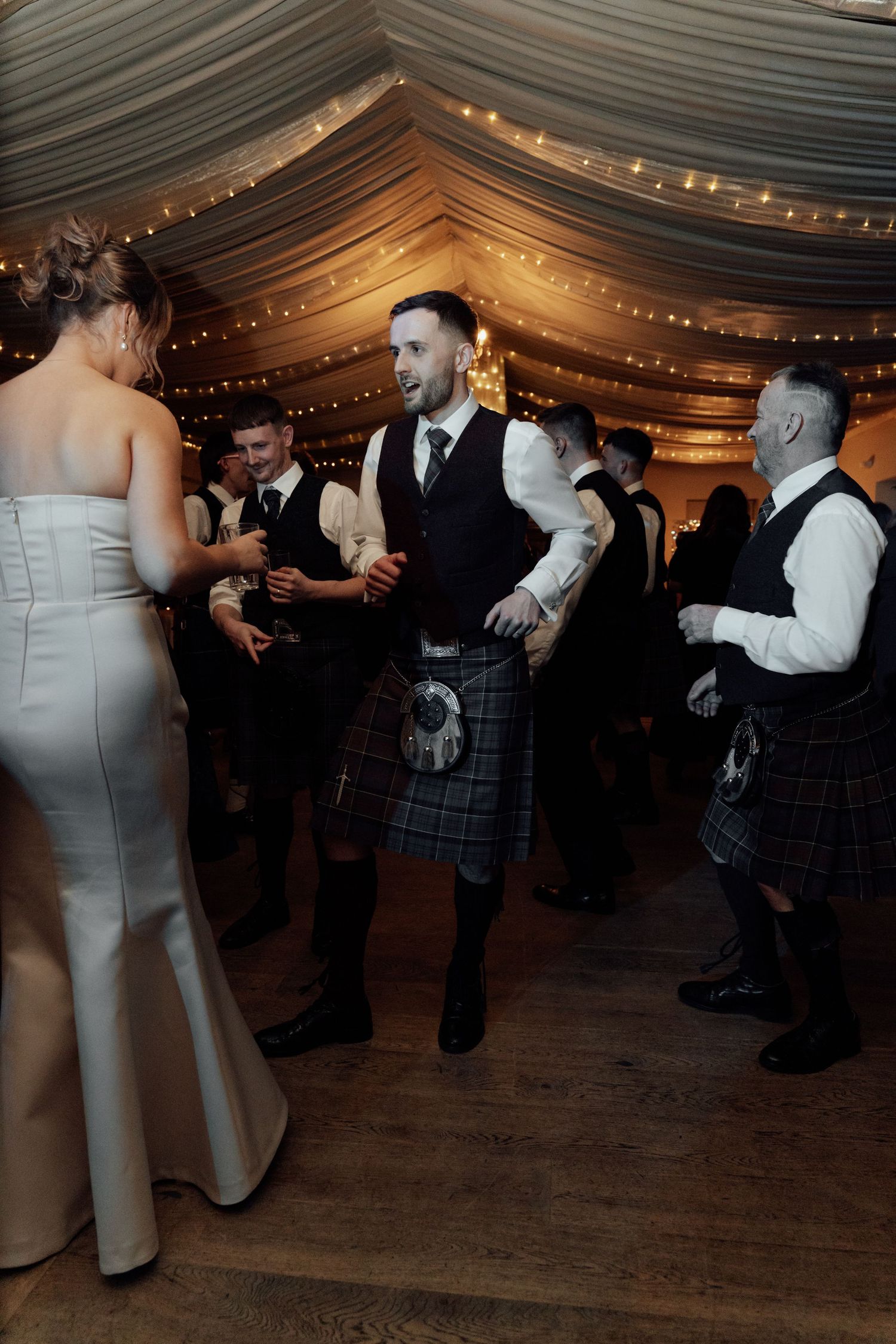 Wedding reception dance floor with Scottish kilts and formal attire under draped ceiling.