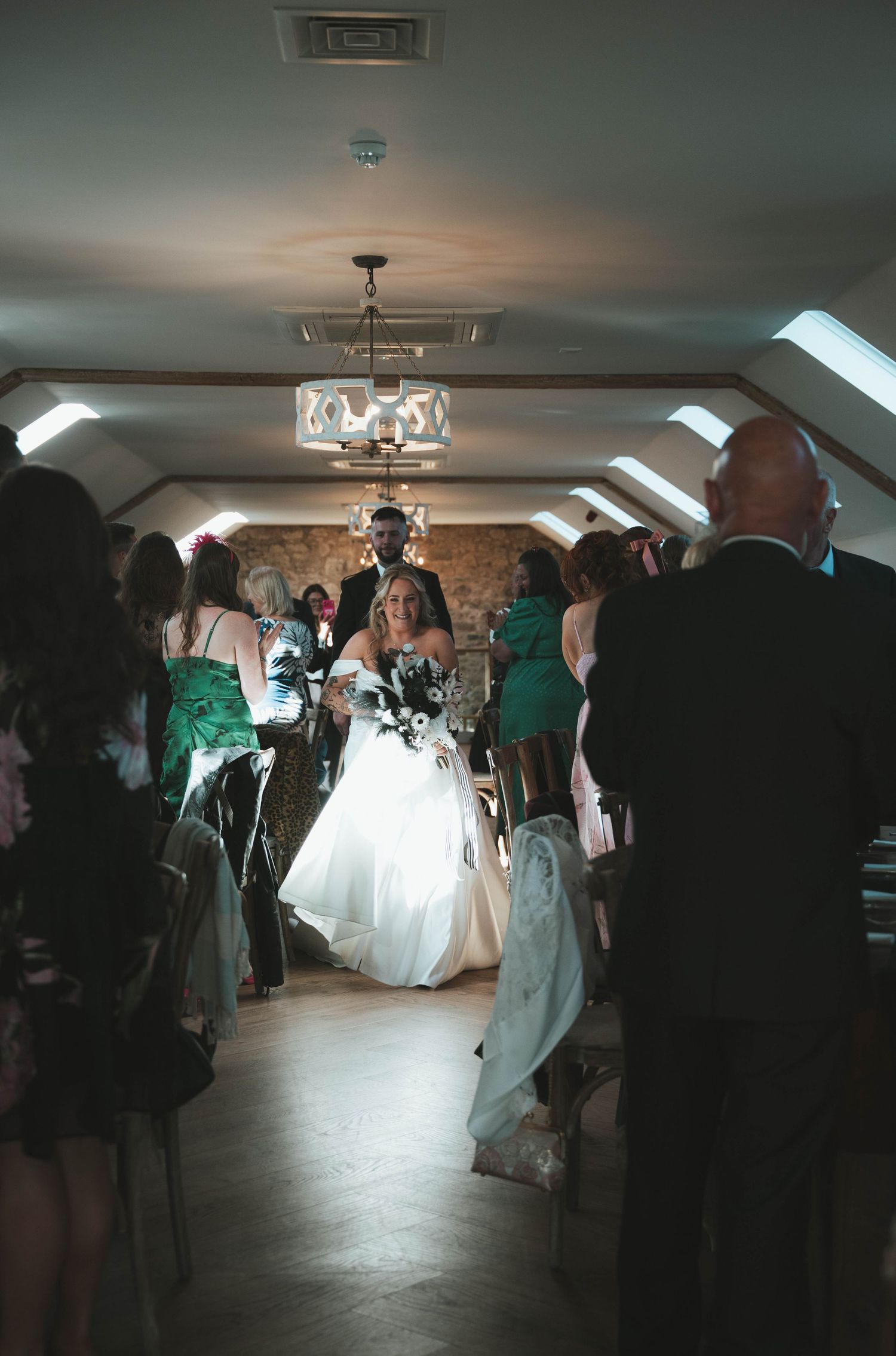 Bride walks down the aisle in a white gown at an indoor wedding ceremony with guests in emerald dresses.