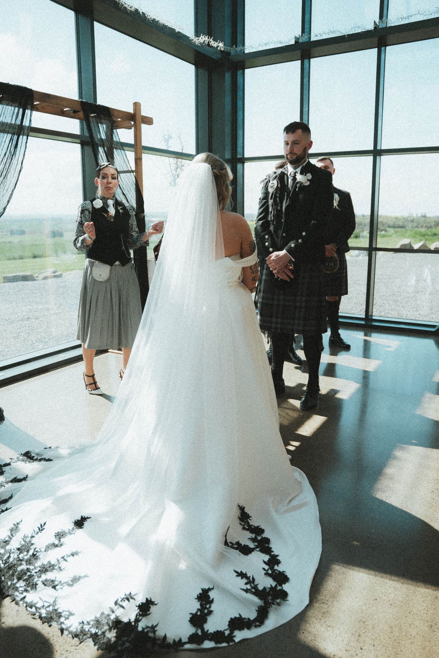Bride in lace-trimmed cathedral veil at altar with groom in Scottish tartan kilt at modern venue.