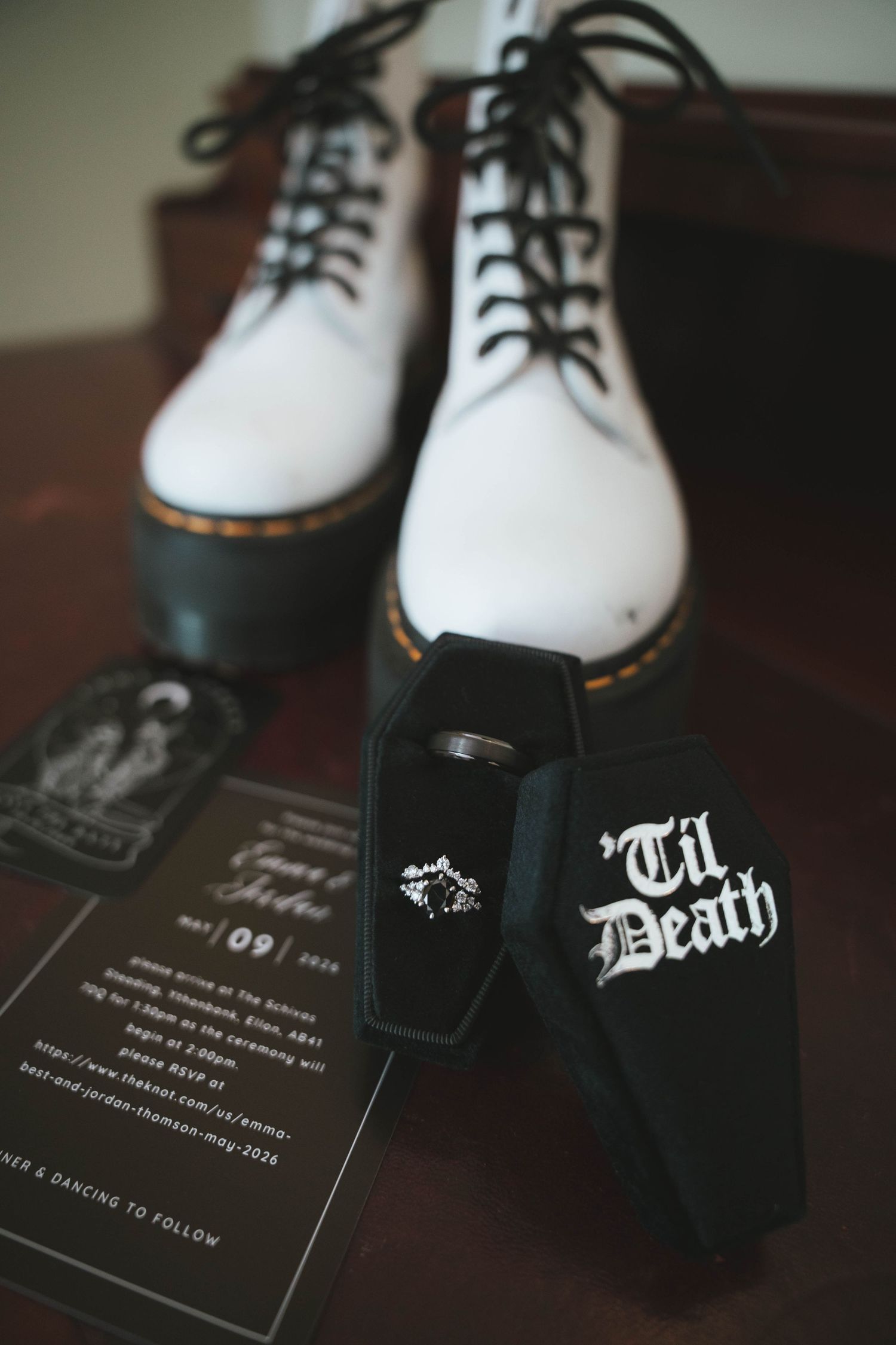 White Doc Martens boots with coffin-shaped Til Death ring box and wedding rings beside dark floral wedding invitation.