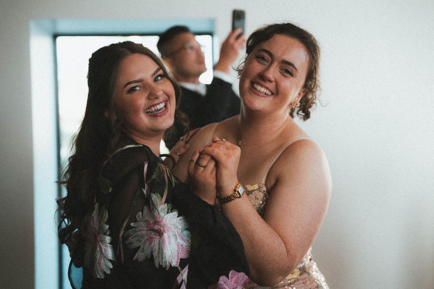 Two laughing bridesmaids in floral dresses hold hands joyfully while man takes selfie behind them.