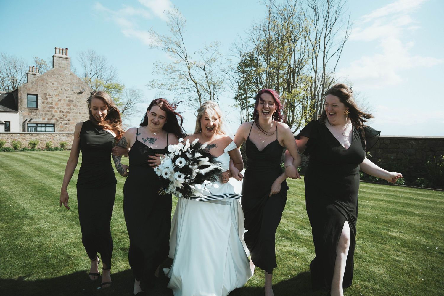 Bride in white gown walking with four bridesmaids in black dresses on sunny lawn.