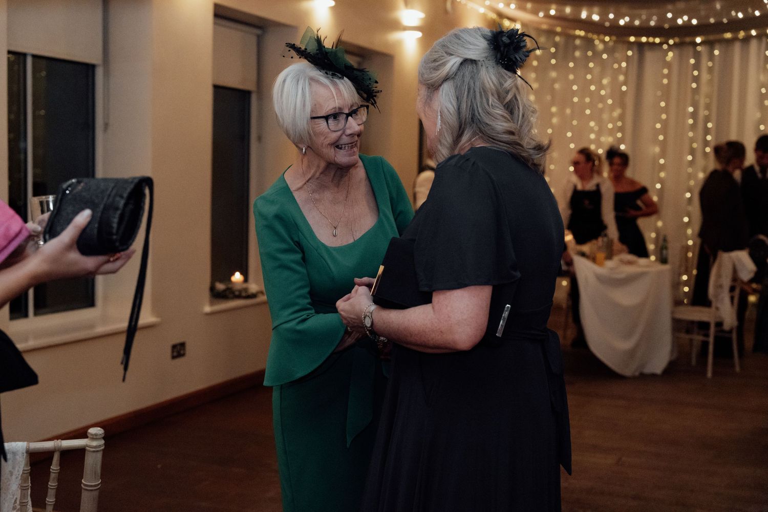 Wedding guests in festive attire chatting in venue with fairy light decorations.