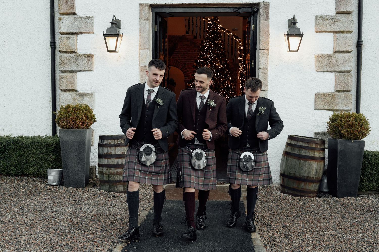 Three men in traditional Scottish kilts walk down steps at formal venue entrance.