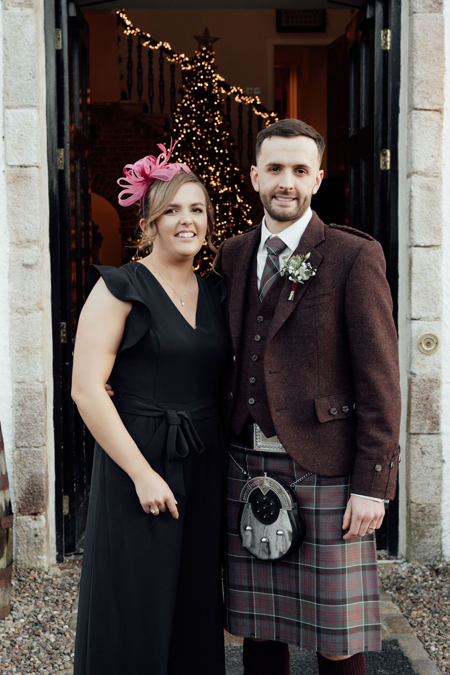 Formal wedding portraits with Scottish kilts and black dresses against stone building entrance.