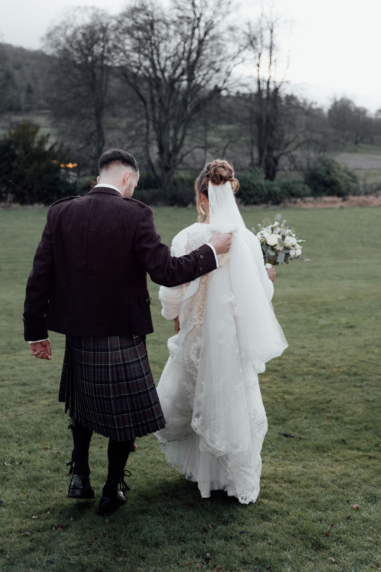 Newlyweds stroll through a grassy field, with the groom wearing a Scottish kilt and bride in a white gown.