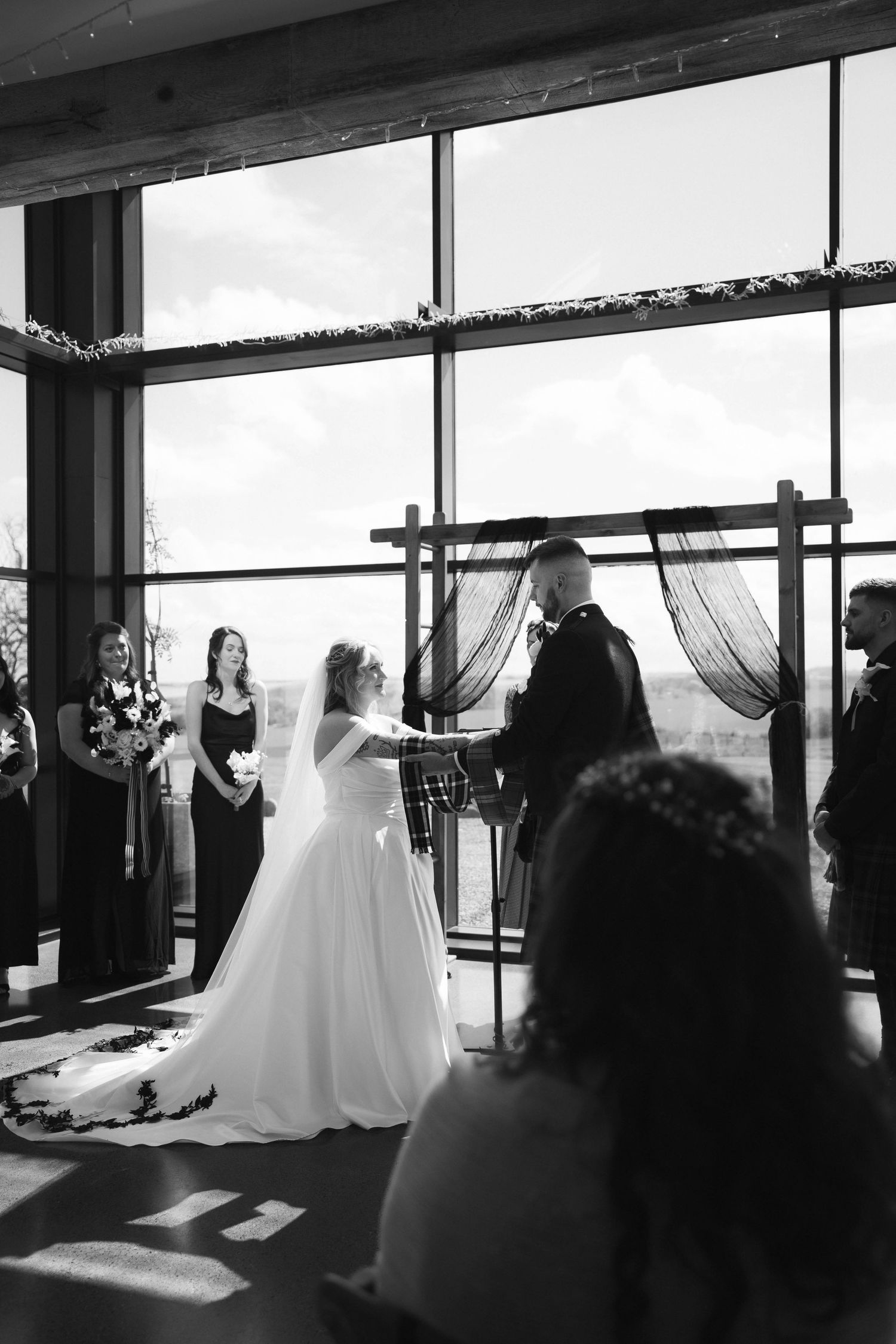 Black and white photo of bride and groom exchanging vows at altar before large windows with wedding guests seated.
