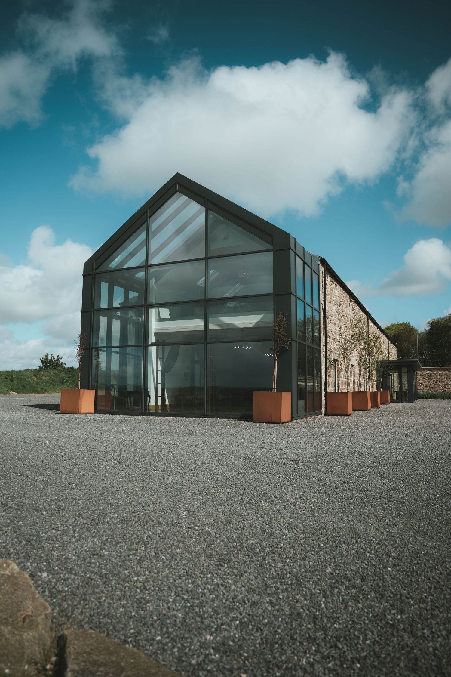 Modern glass and steel barn-style building with copper planters on a gravel forecourt under a blue cloudy sky.