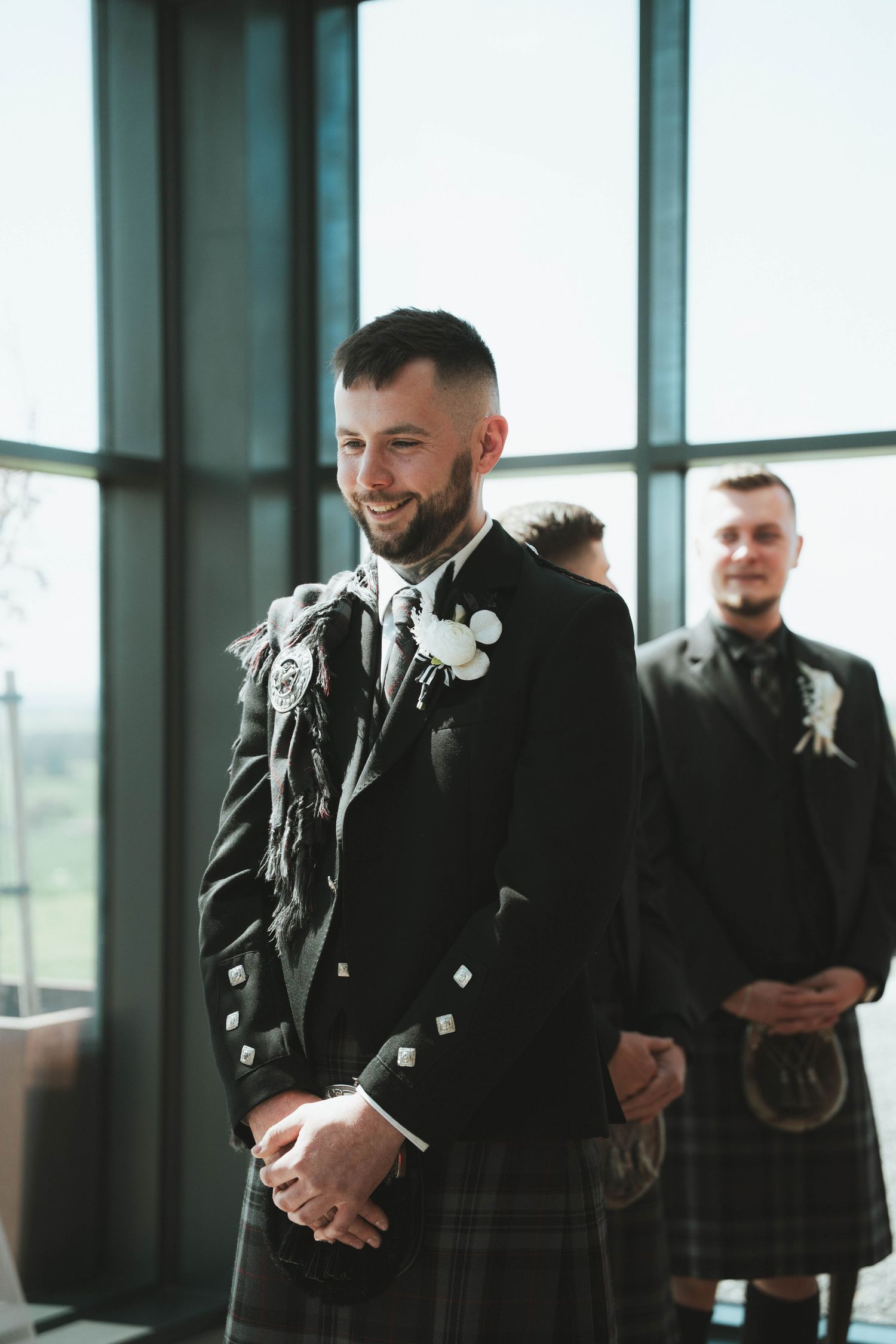 Smiling groom in traditional Scottish black kilt outfit with boutonniere stands at wedding ceremony altar.