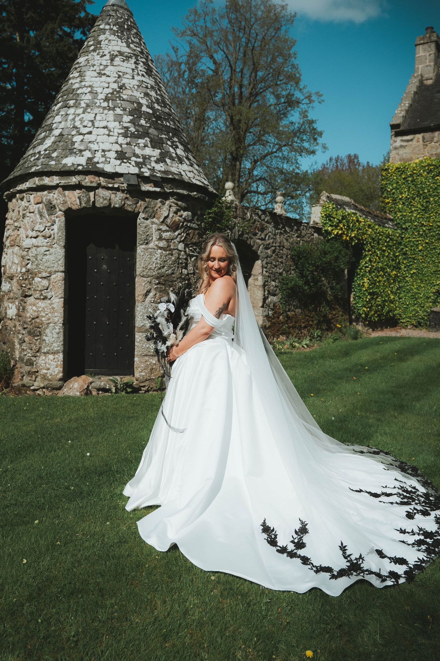 Bride in stunning white gown with black floral train twirls before historic Scottish stone tower.