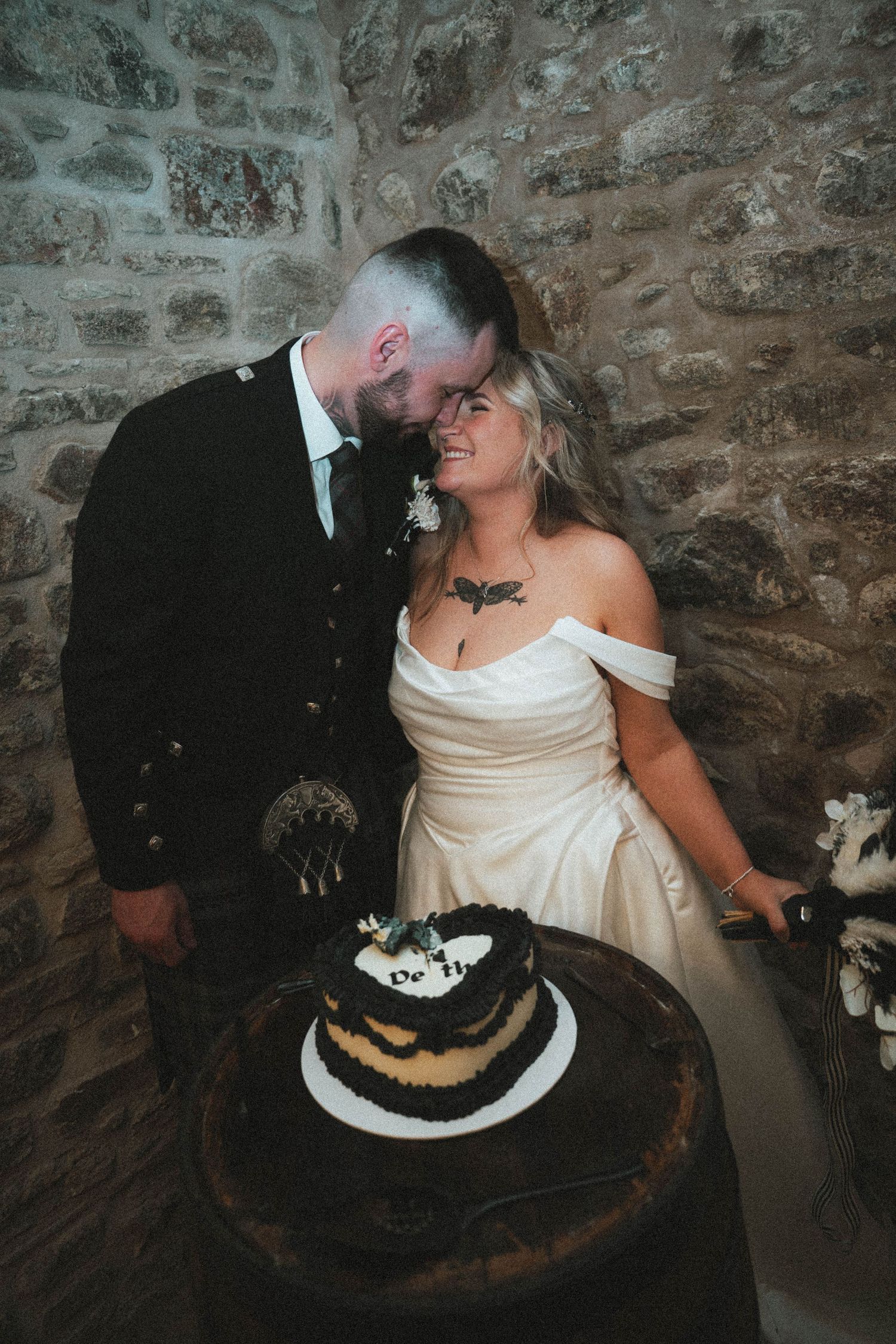Newlywed couple smiling together while cutting small decorated wedding cake on barrel against rustic stone wall backdrop.