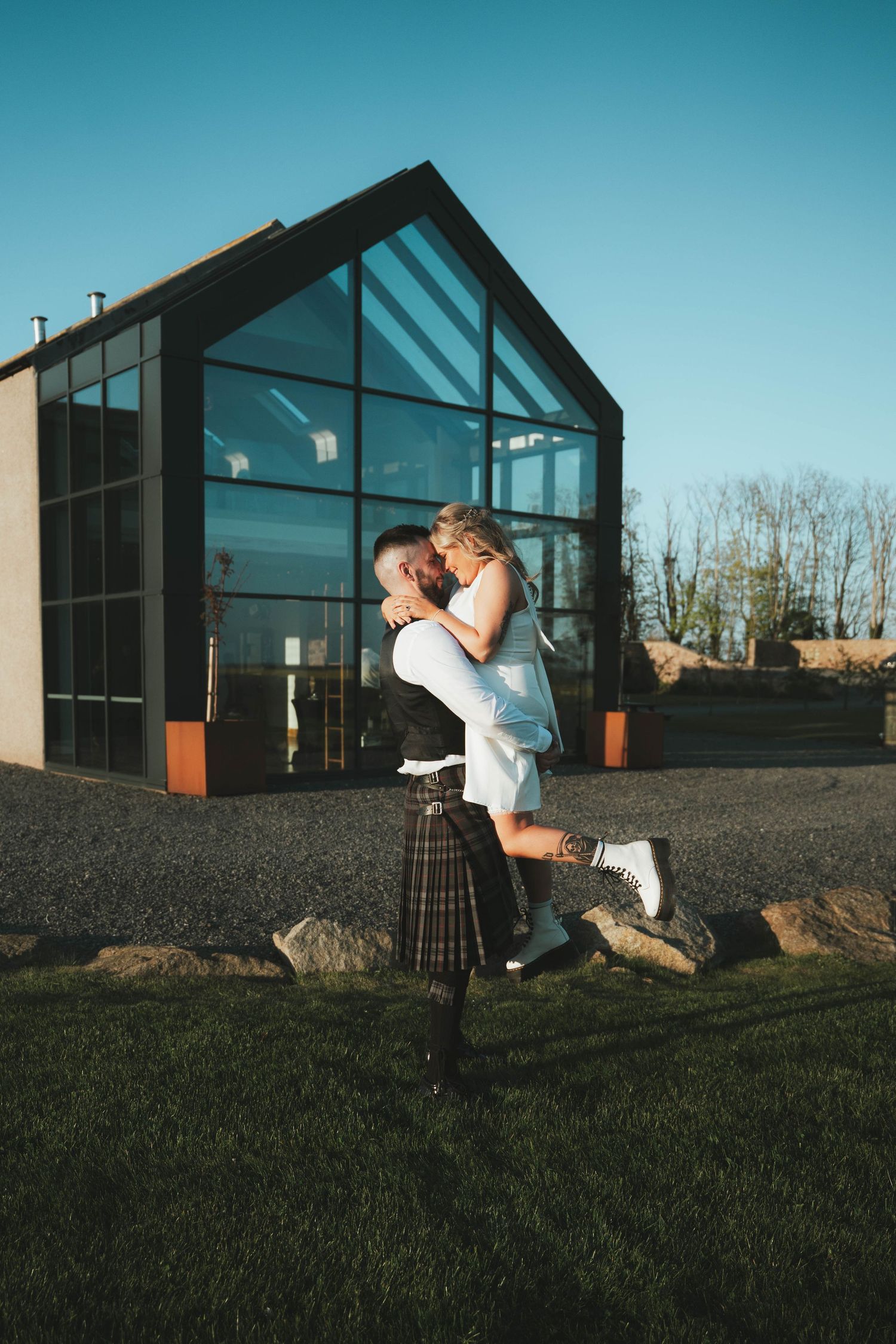 A man in a kilt lifts his bride outside a modern glass building on their Scottish wedding day.