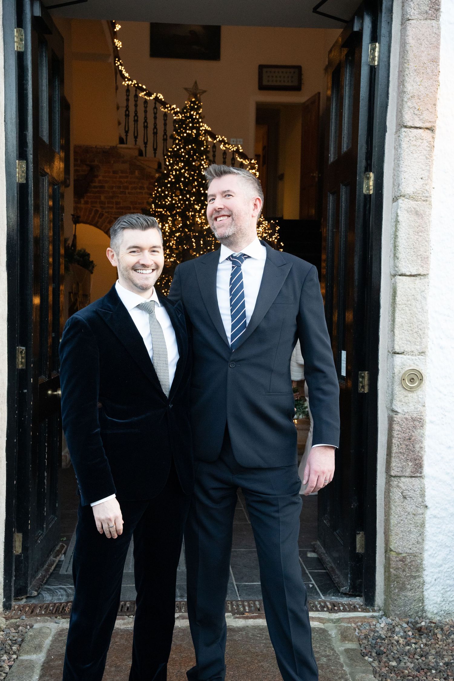 Groomsmen in black suits and ties share a candid moment outside a venue decorated with holiday lights.