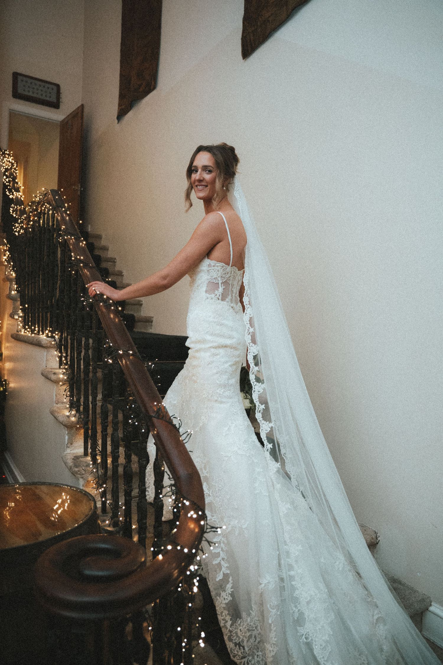 Bride twirls in her lace wedding gown beside a decorated staircase with twinkling lights.