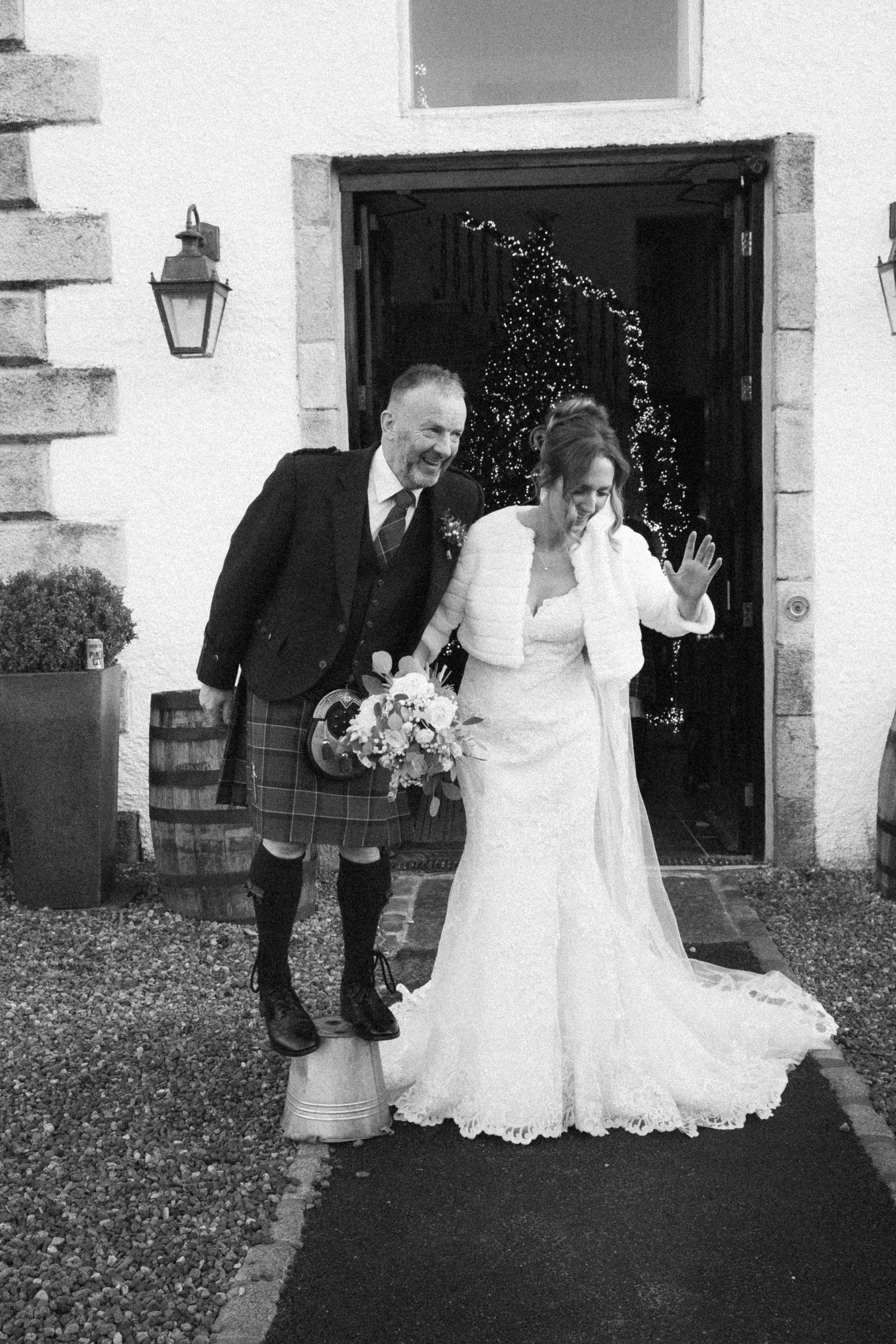 A black and white wedding photo showing a joyful moment outside a venue with fairy lights.