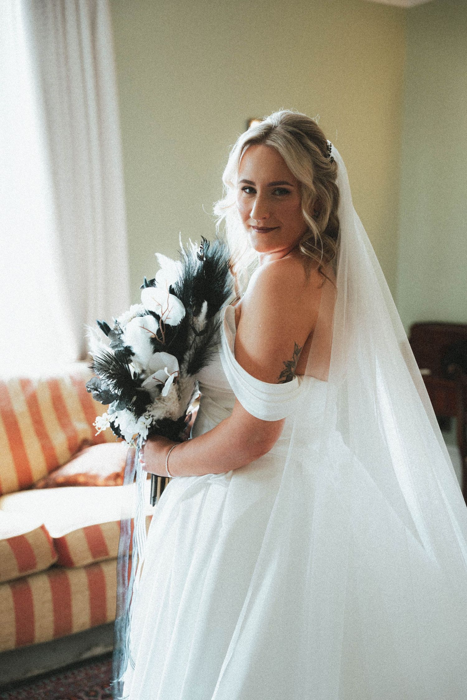 Smiling bride in white off-shoulder gown holding black and white bouquet with veil.