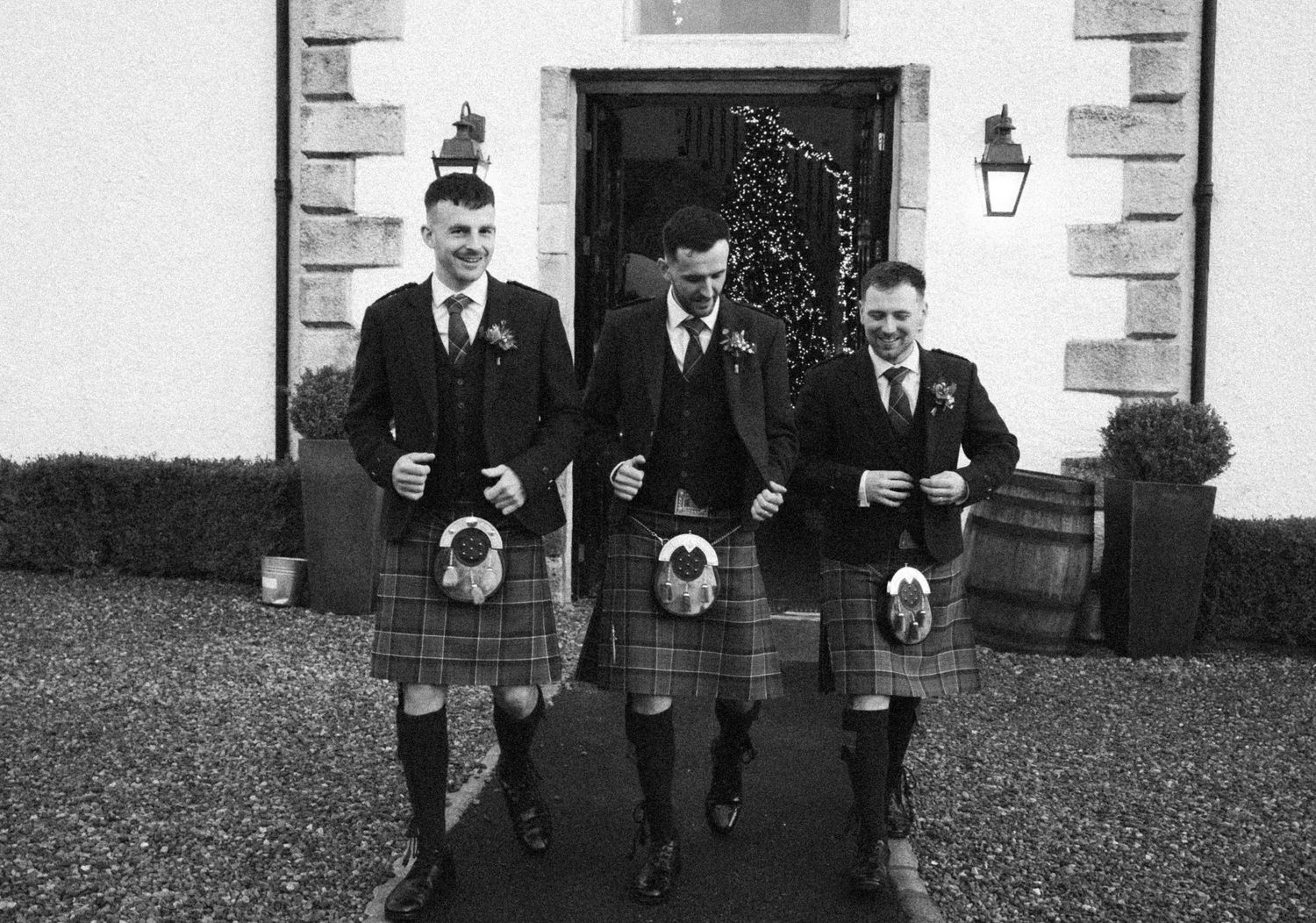 Black and white image of three men in kilts walking together at venue entrance.