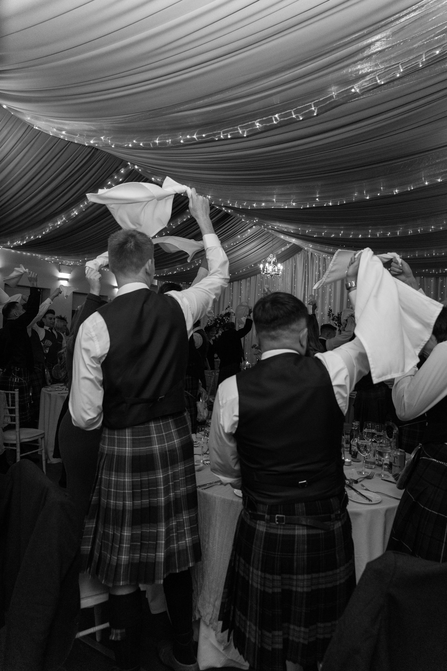 Wedding guests in traditional Scottish kilts dancing together under draped ceiling with fairy lights.