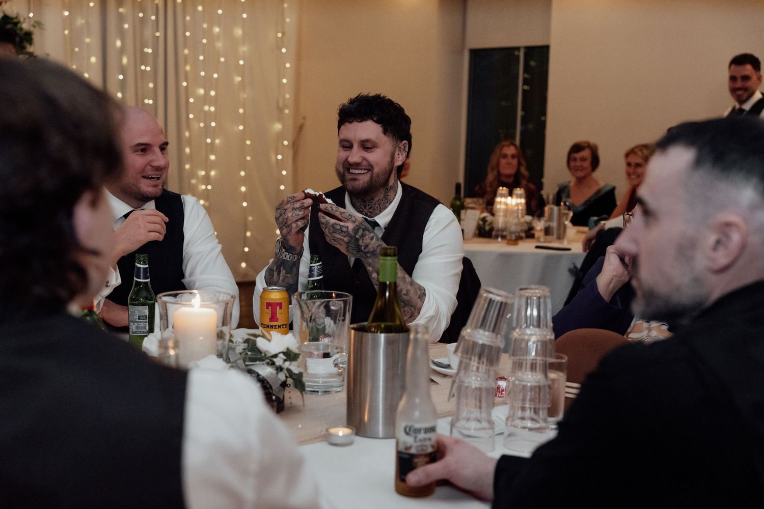 Wedding guests enjoy conversation and drinks at a reception table decorated with candlelight and string lights.