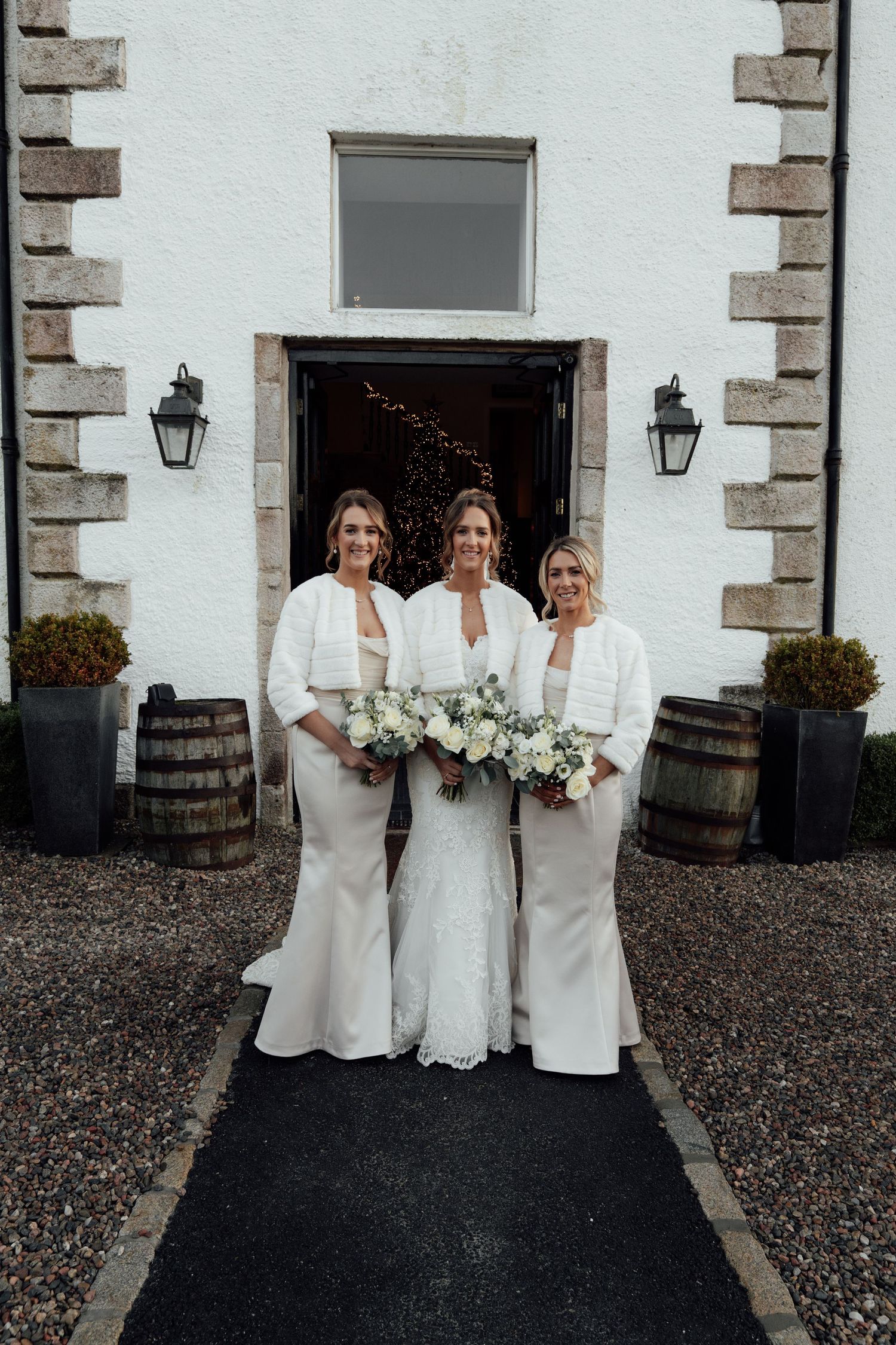 Wedding attendants in white dresses pose with bouquets outside stone building entrance.