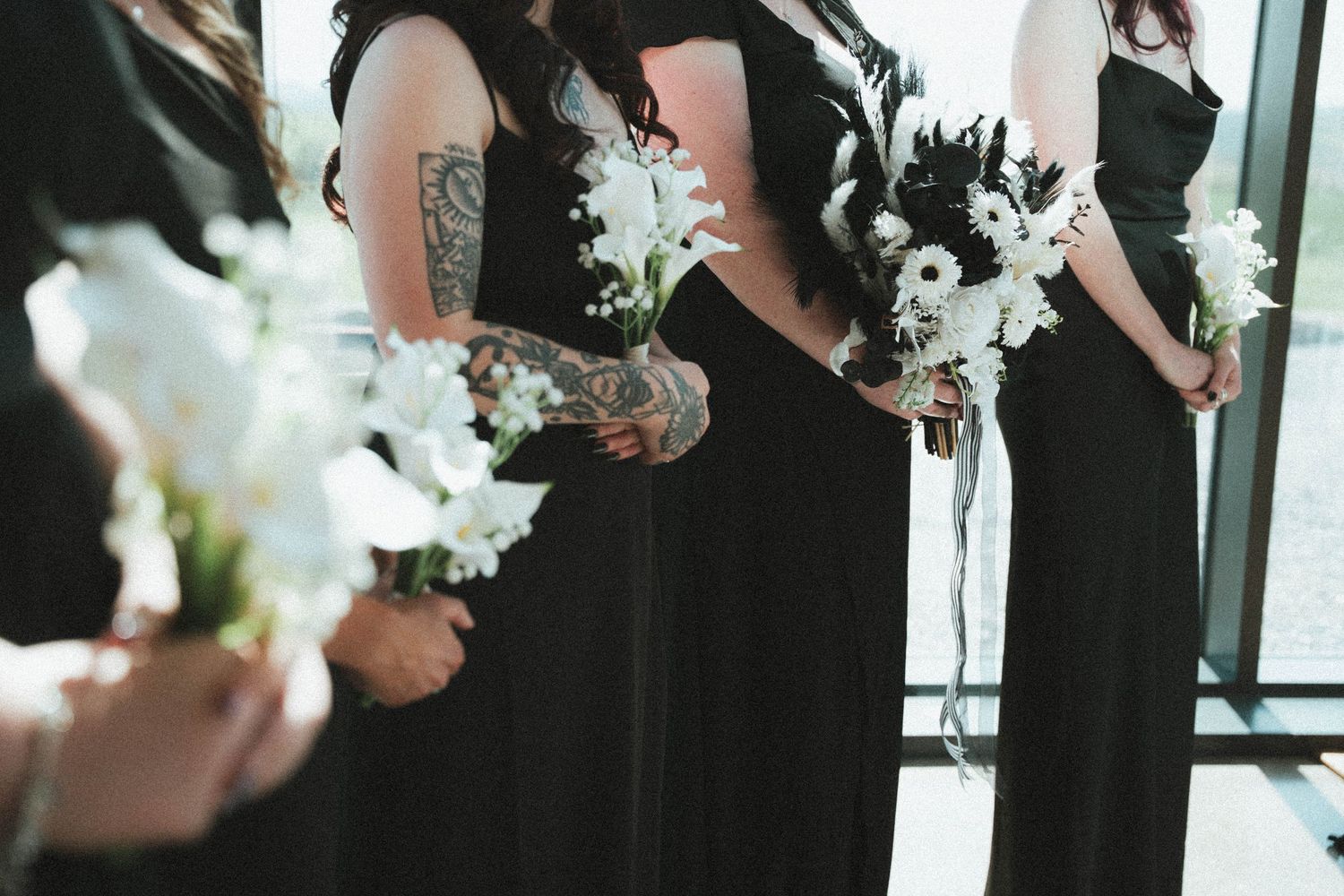 Bridesmaids in black dresses holding white and black floral bouquets, one with a visible arm tattoo.