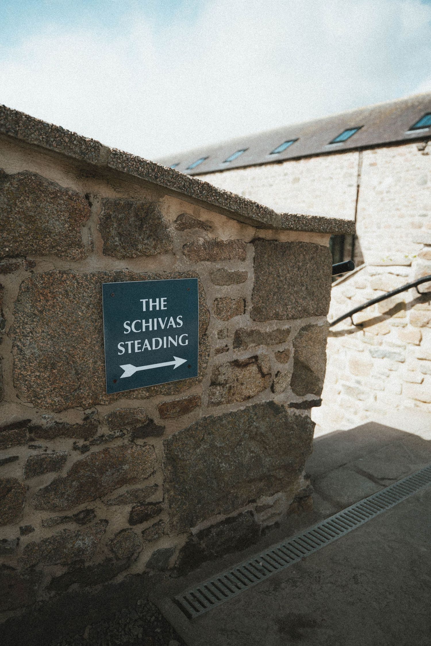 Stone wall with a teal directional sign pointing to The Schivas Steading at a historic Scottish stone building.