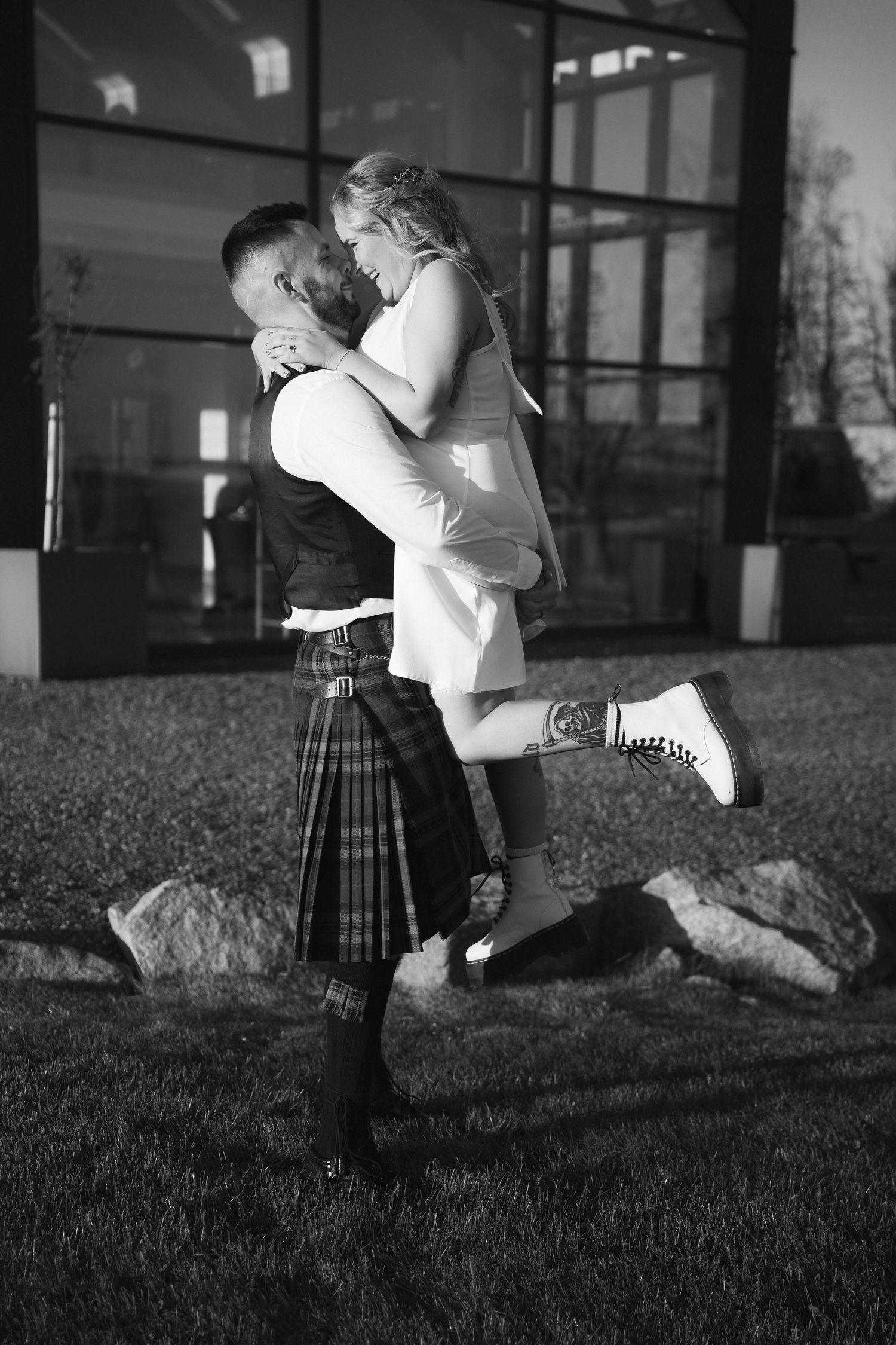 Groom in tartan kilt lifting bride in white dress and Doc Martens in black and white photo.