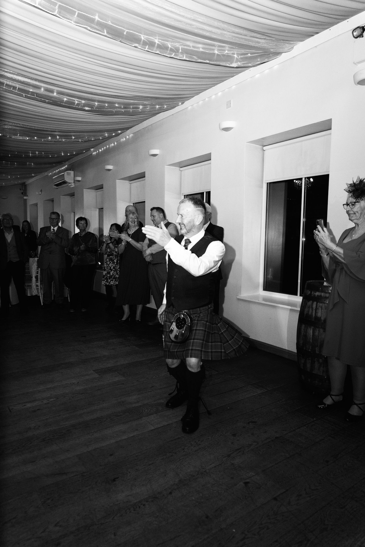 Wedding guests dance in formation at a reception with draped white ceiling and hardwood floors.