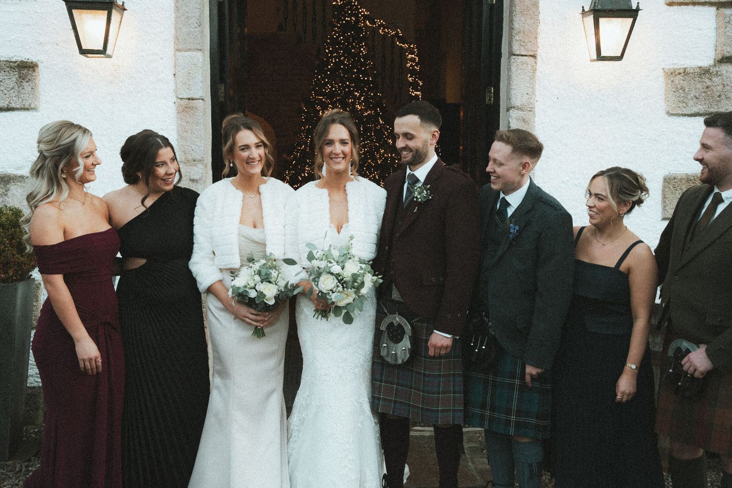 Wedding party members in burgundy dresses and dark suits pose together outside venue entrance.