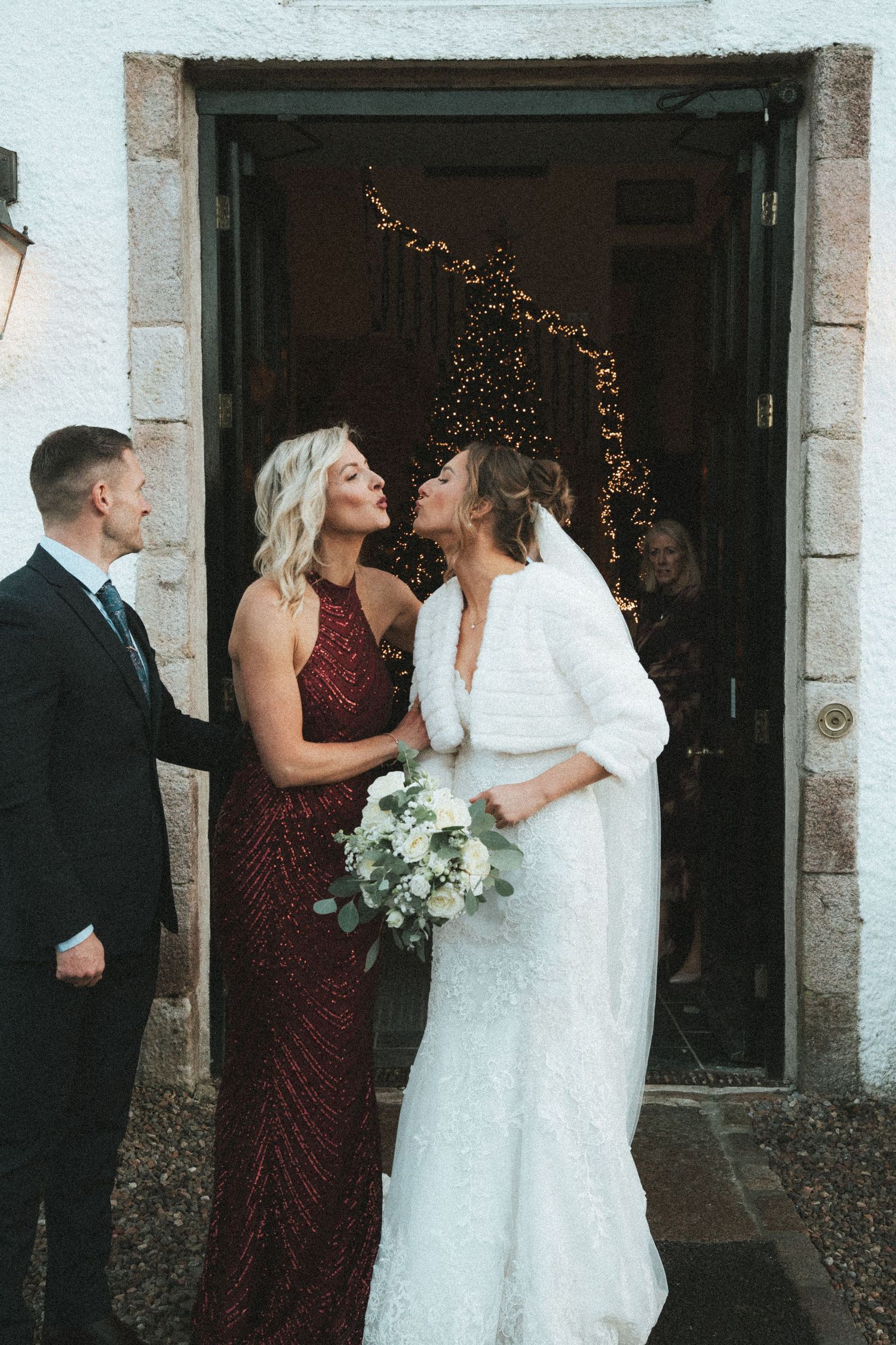 Wedding party members share a joyful moment outside a stone building with white walls and lanterns.