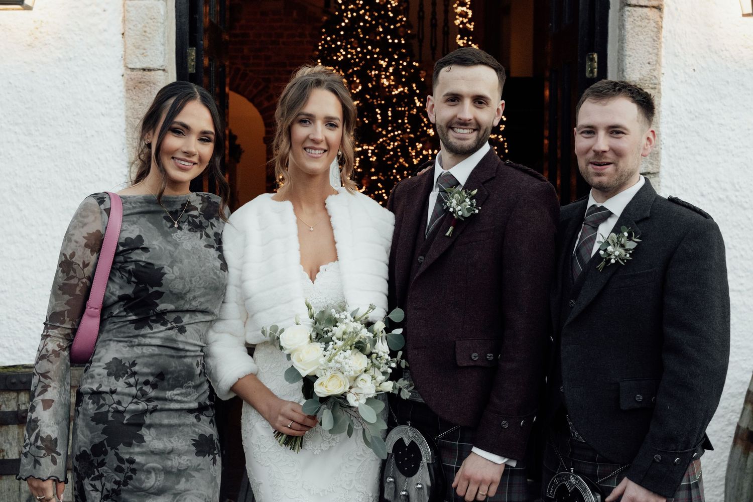 Wedding group poses together in Scottish formal wear outside white stone venue.