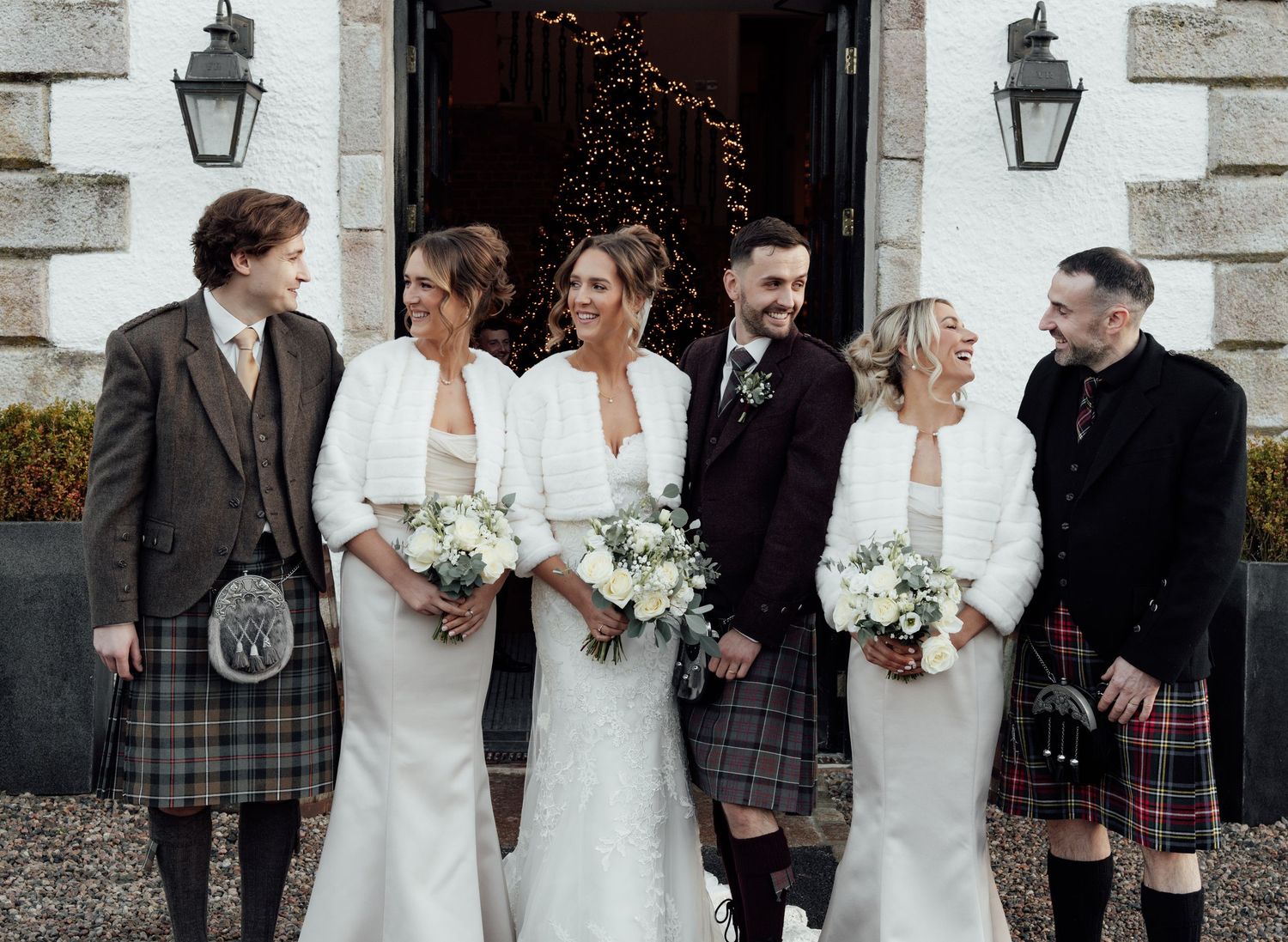 Formal wedding group photo featuring Scottish kilts and white dresses against stone building backdrop.